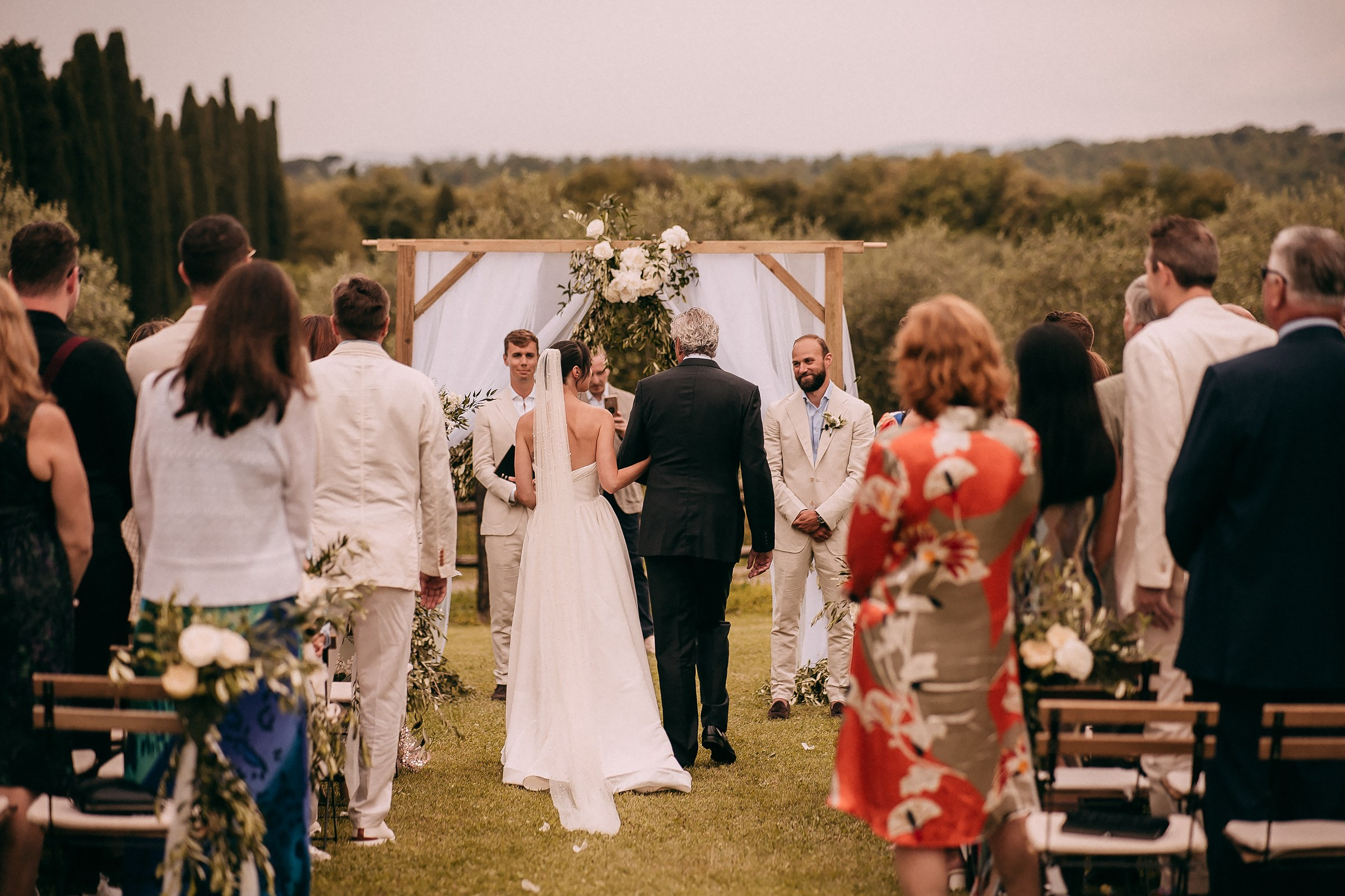 The bride, escorted by her father, approaches the altar under a floral arch, surrounded by guests in elegant attire.