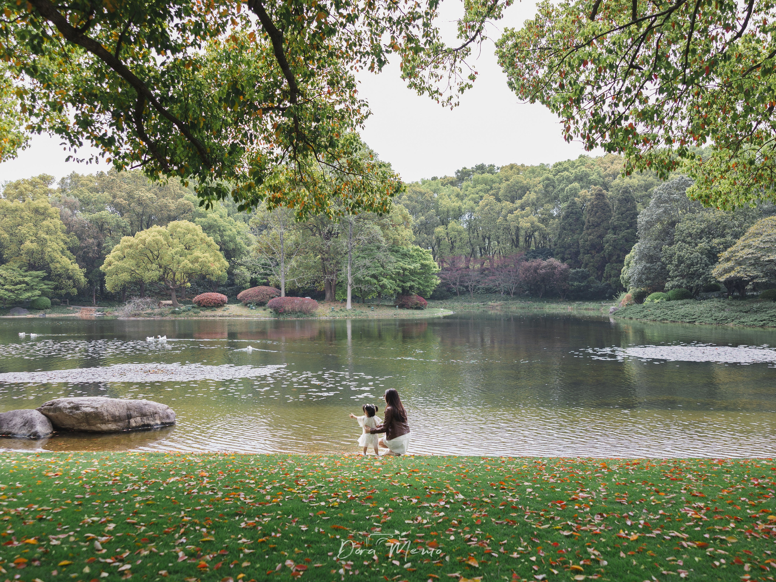 A mother crouches beside her toddler daughter by a lake, pointing at the water together