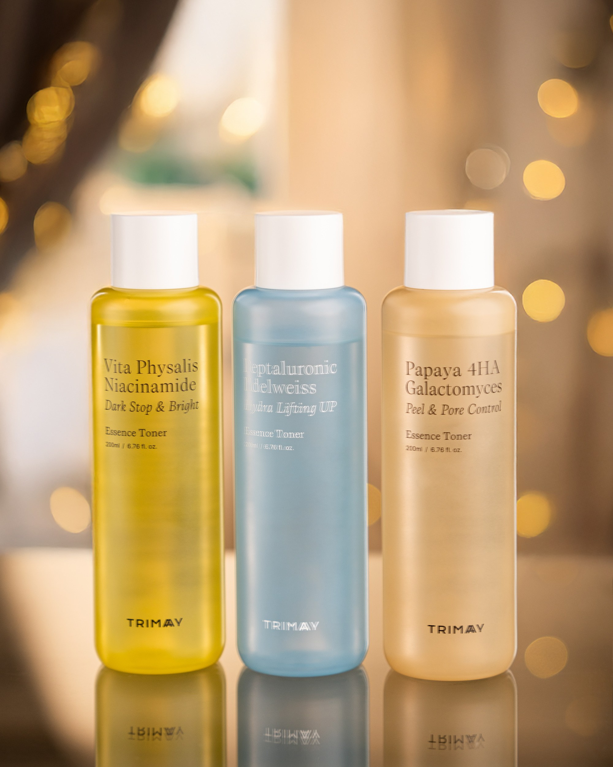 Qızılı və bulanıq işıqlar fonunda, güzgü effekti verən səth üzərində, Trimay markalı üç fərqli rəngdə Essence Toner butulkası: Vita Physalis Niacinamide (sarı), Heptaluronic Edelweiss (mavi) və Papaya 4HA Galactomyces (bej).