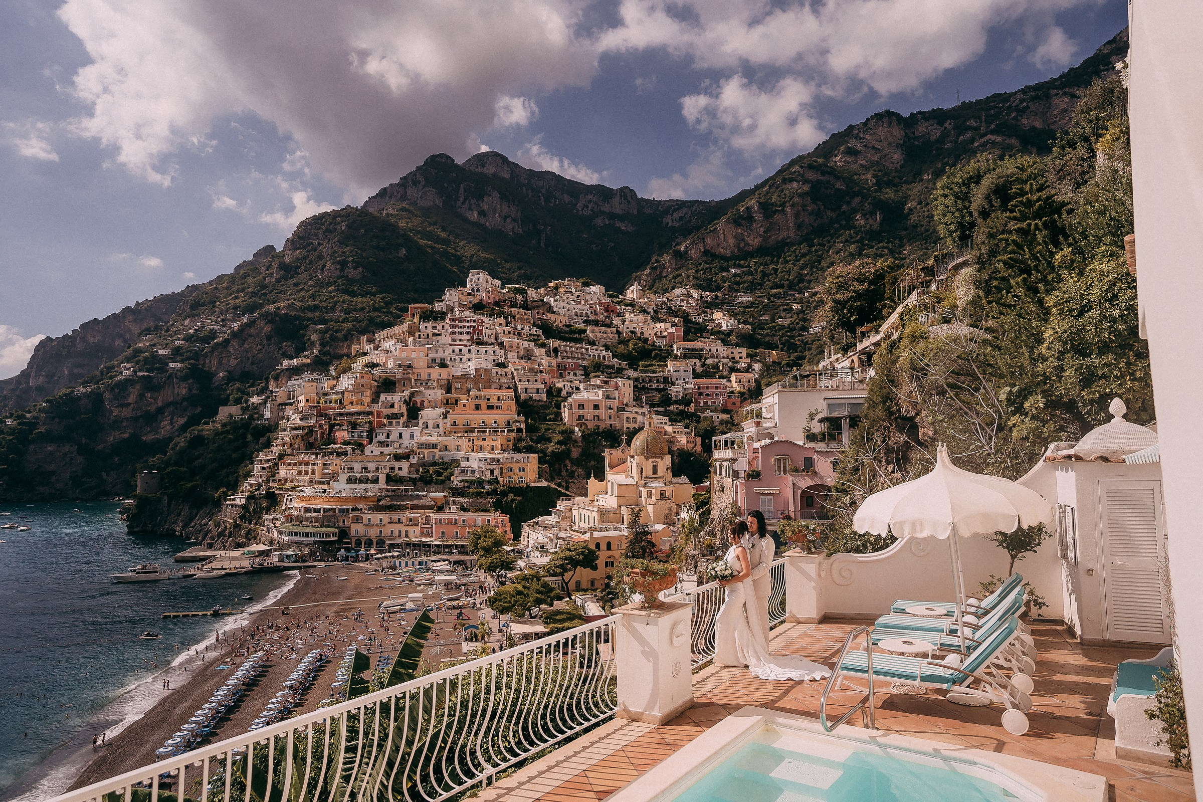 A wide-angle view of the villa’s terrace showcasing the newlyweds amidst Positano’s breathtaking landscape, with lounge chairs and a pool completing the idyllic scene.