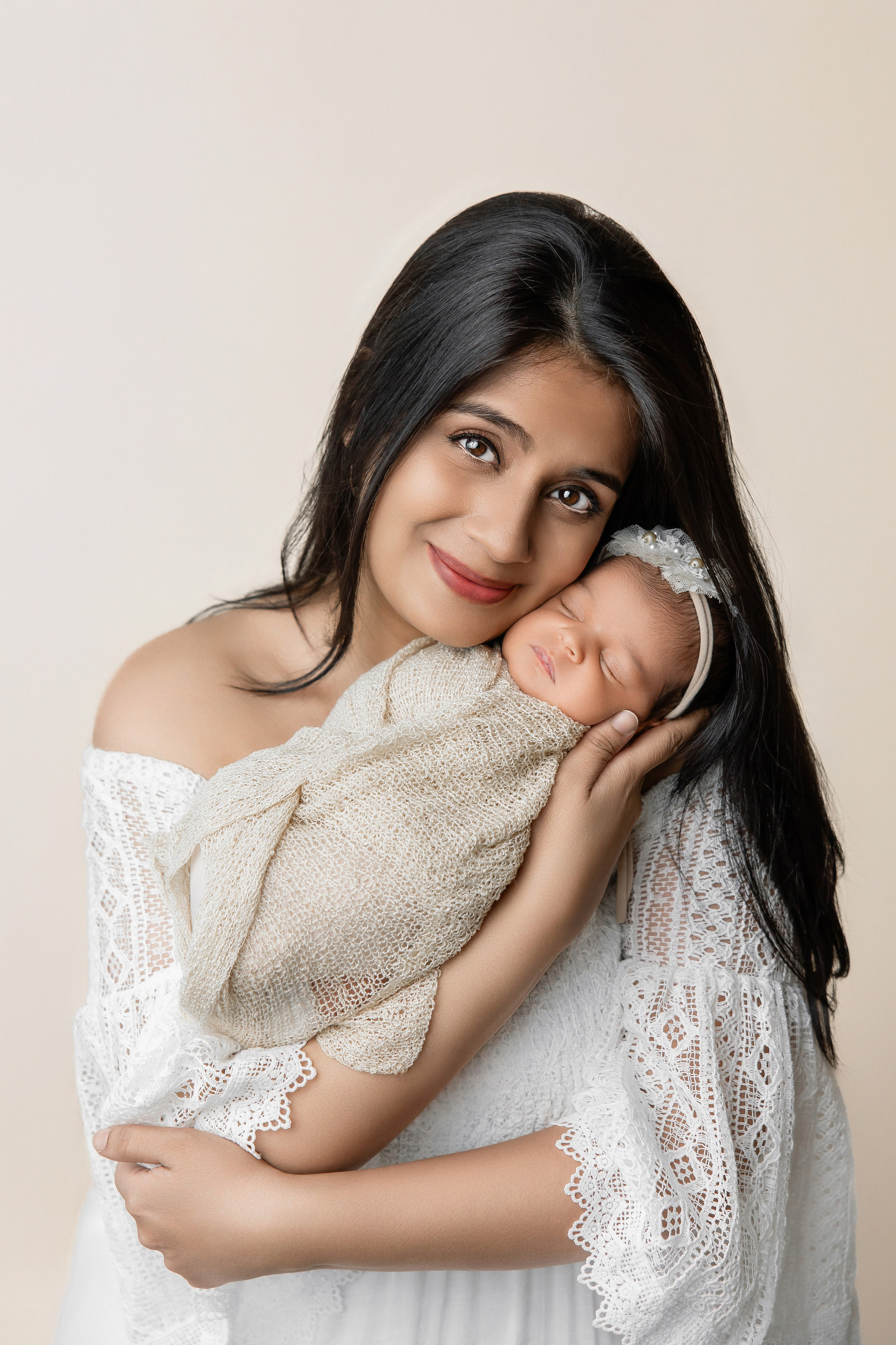 Smiling mother in a white lace dress holding her newborn baby wrapped in a beige blanket — professional studio portrait with soft, natural tones.