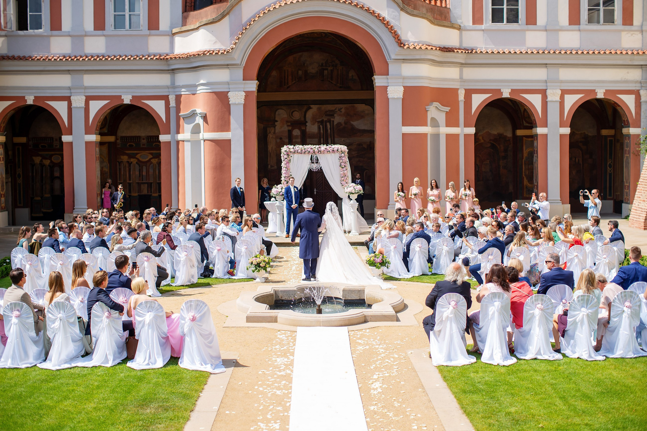 The father of the bride wearing a gray top hat escorts his daughter to the waiting groom as several hundred seated guests look on from their seats at the historic Ledebour Garden.