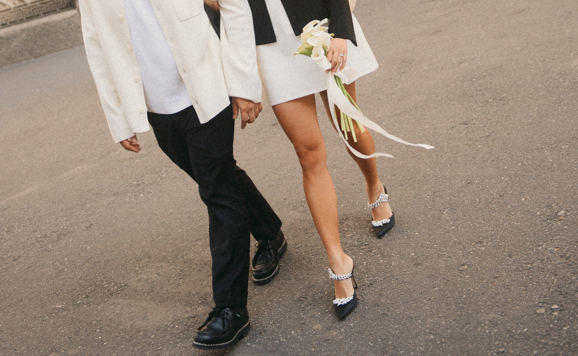 Elegant and modern wedding photo of a bride and groom walking hand in hand, featuring refined shoes and a white calla lily bouquet. A dynamic and intimate image that captures the beauty of movement, love, and the beginning of a shared journey. wedding photography, bride and groom walking, calla lily bouquet, wedding details, modern wedding, editorial wedding photography, elegant wedding, wedding couple, emotional wedding photo, destination wedding