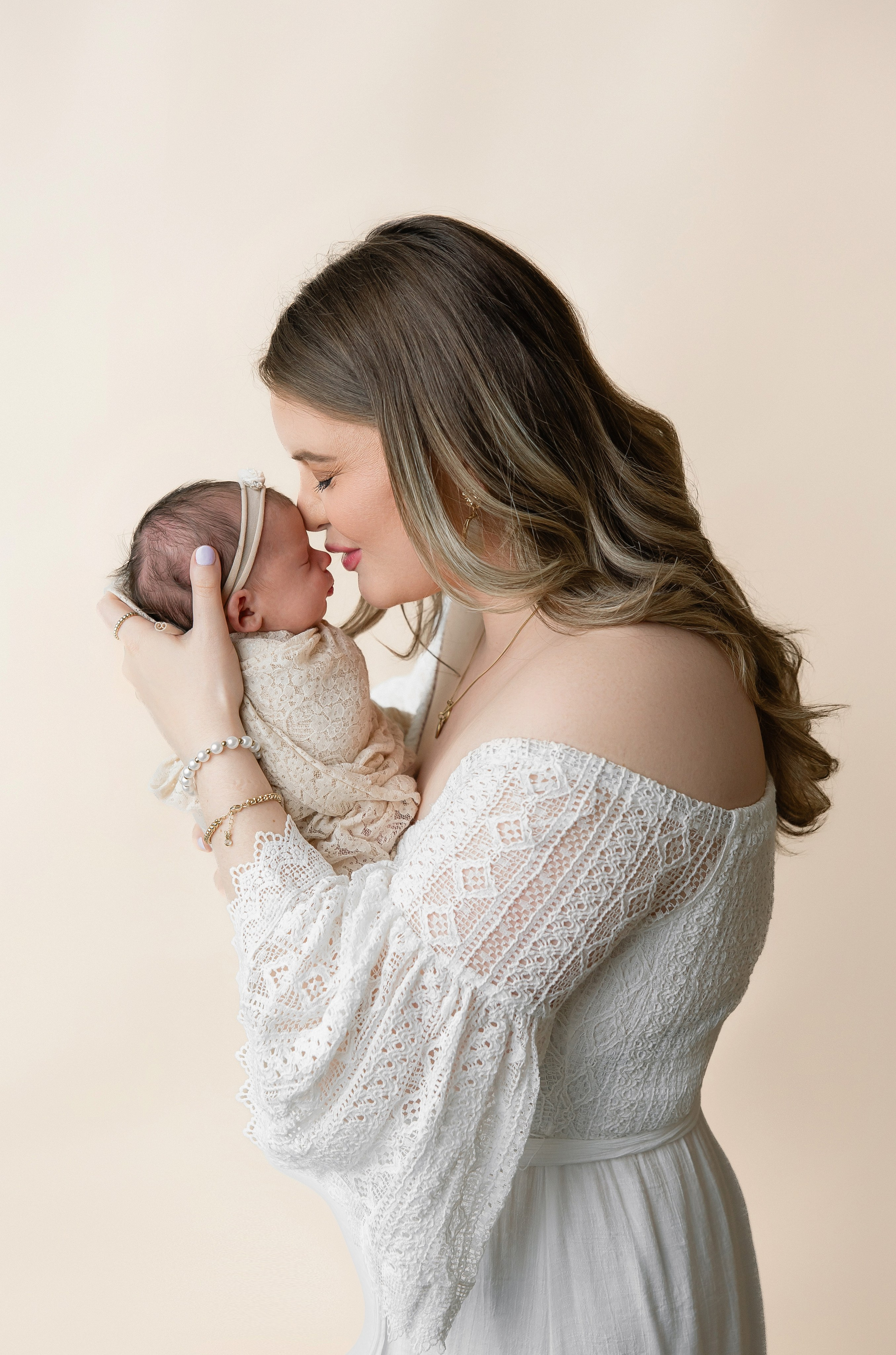 Mother in a white lace dress holding and smiling at her newborn baby dressed in a beige outfit — soft and intimate mother and baby portrait in studio setting.