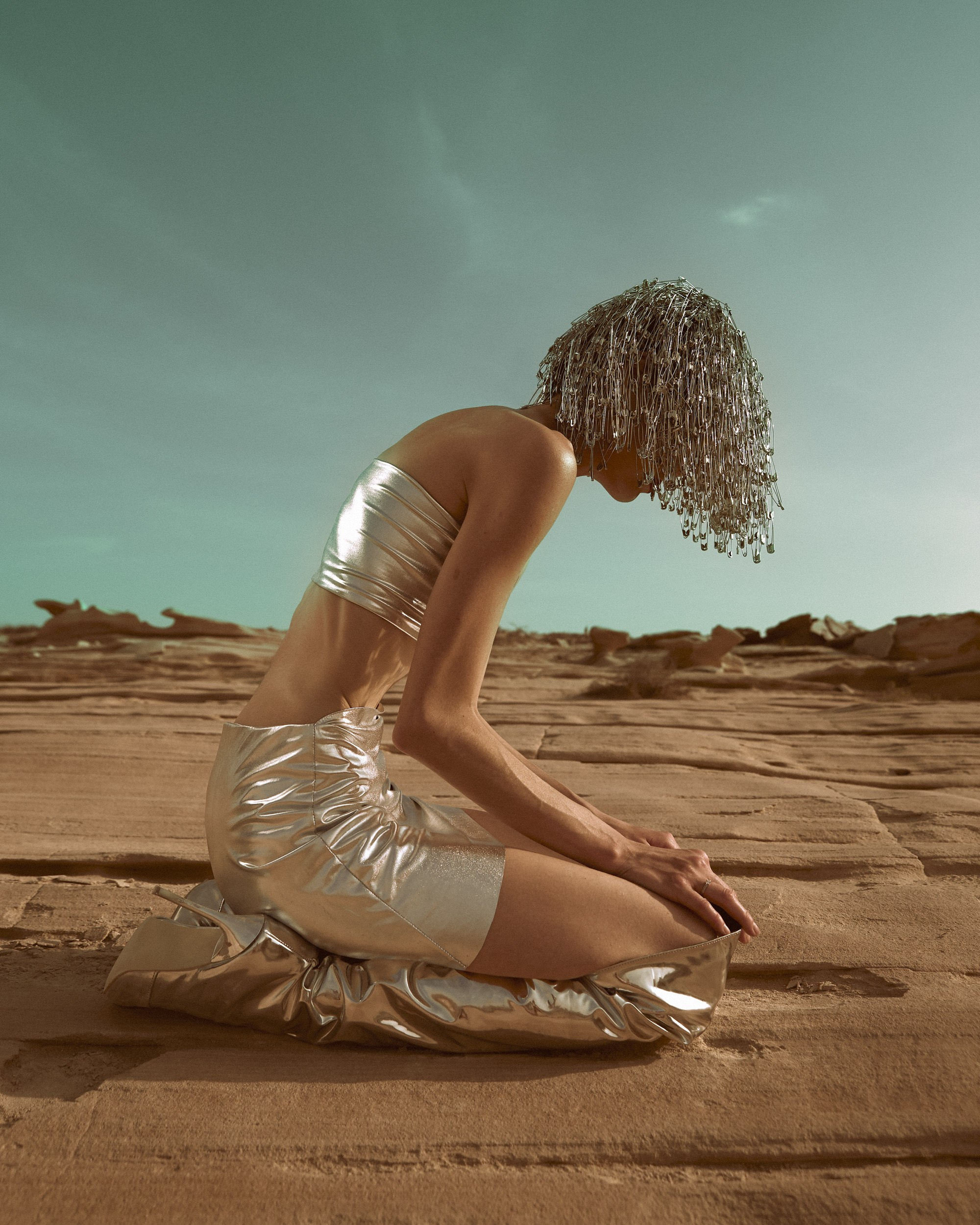 Avant-garde photoshoot featuring a woman in a shiny silver costume, kneeling in a rocky desert setting