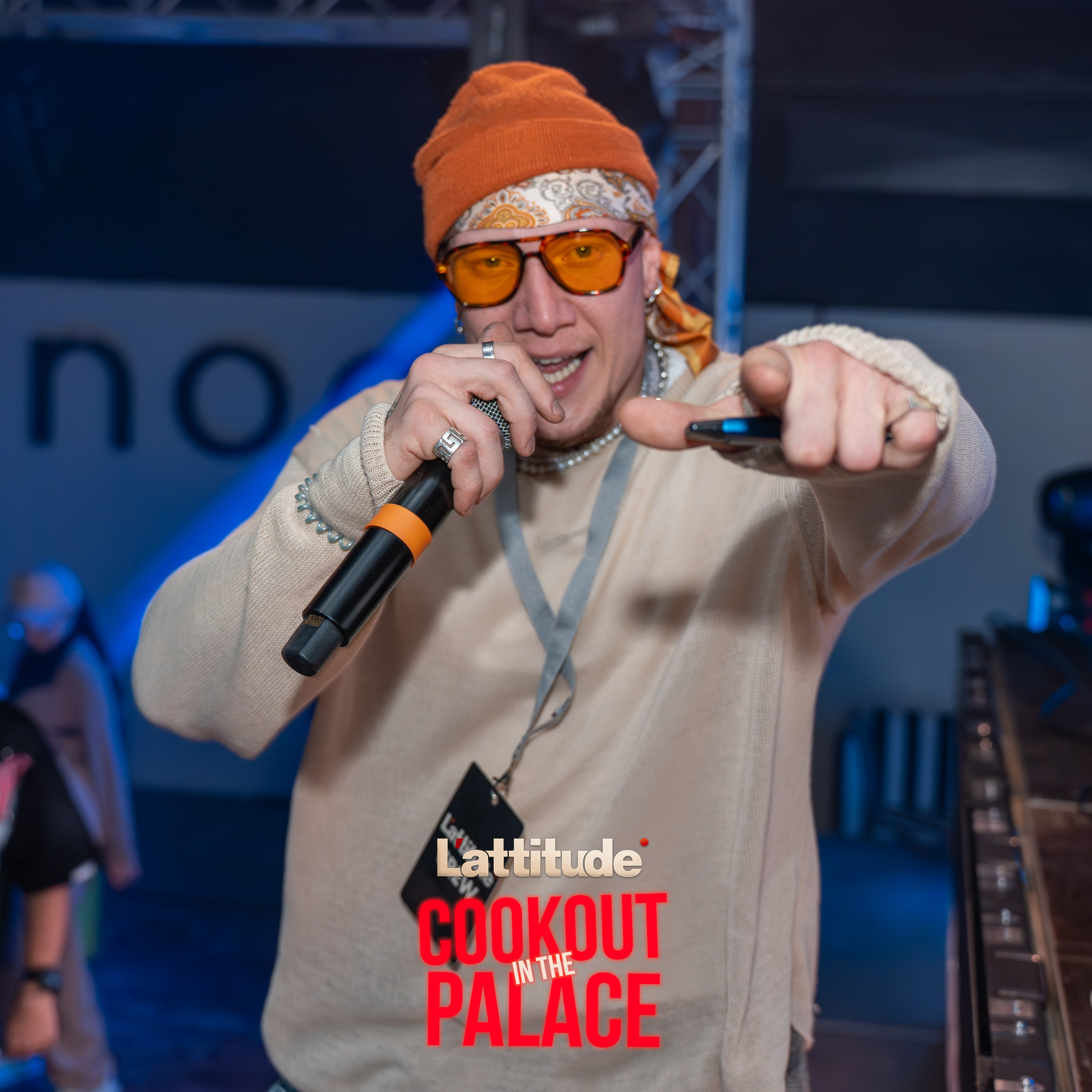 Man wearing orange beanie and tinted glasses holding a microphone, pointing at the camera at an event.