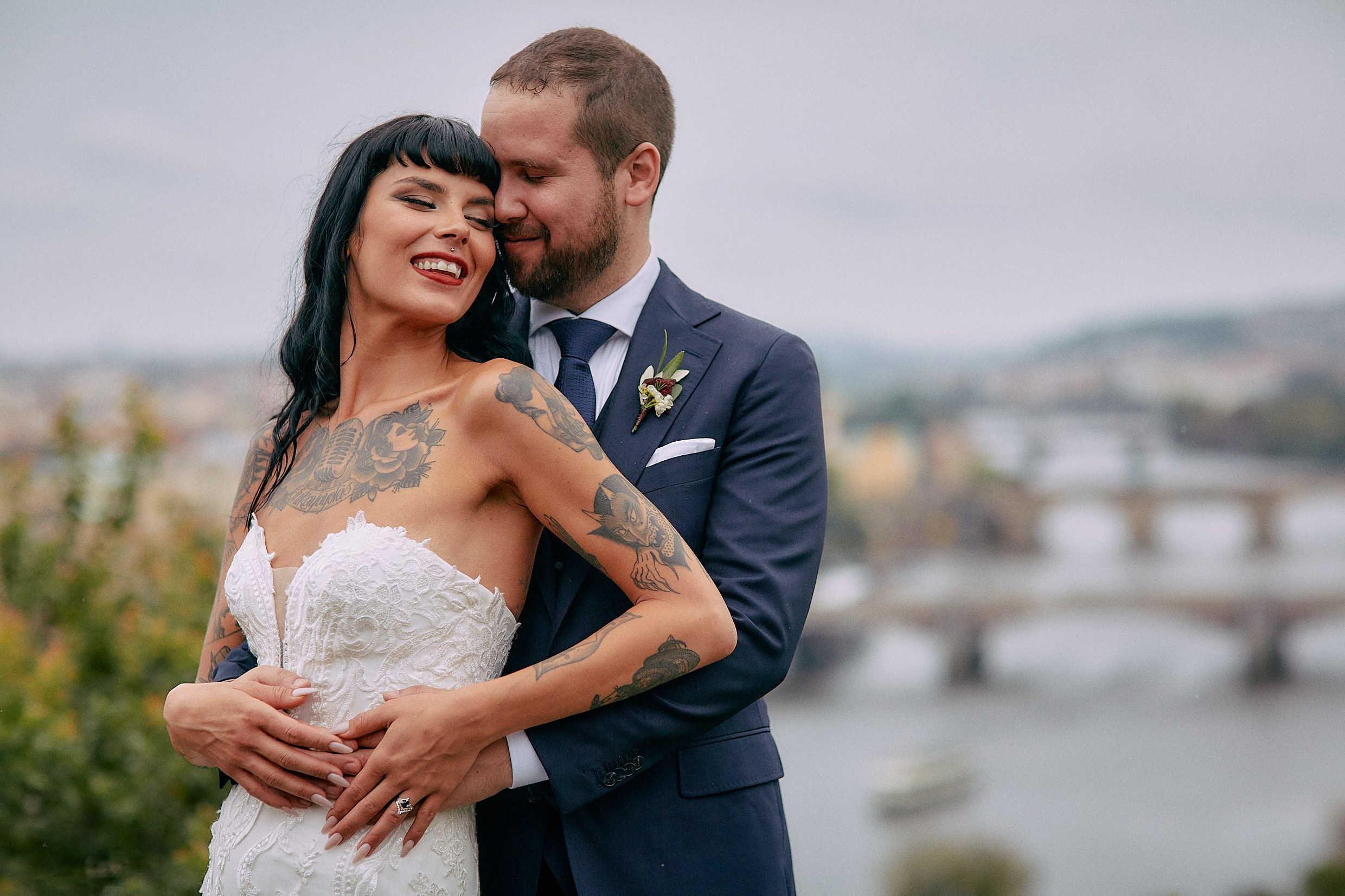 Groom embracing bride from behind at rainy Letná viewpoint.