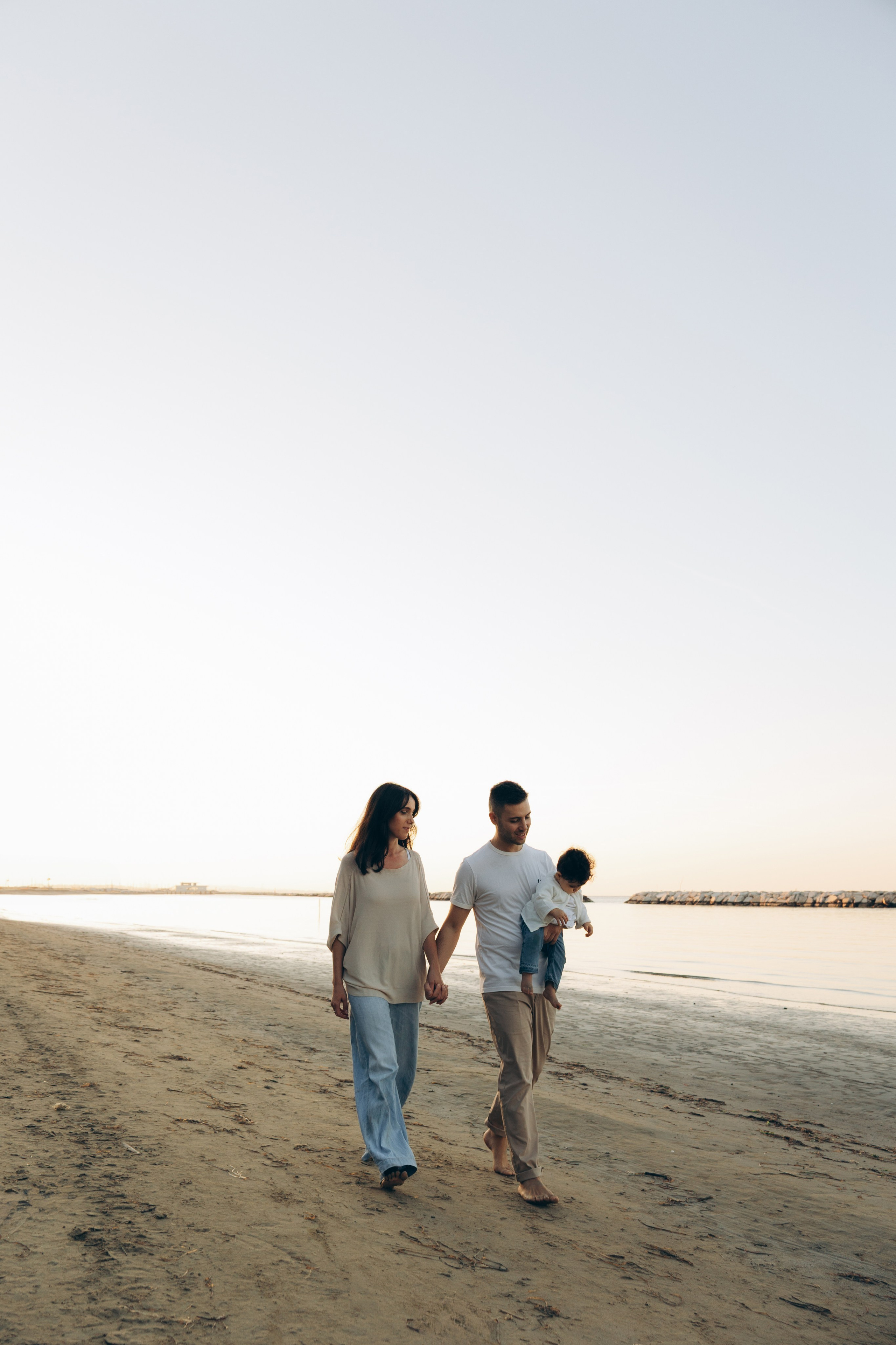 Servizio fotografico di famiglia al mare a Cattolica: madre e padre camminano a piedi nudi sulla spiaggia lungo il mare Adriatico, il padre tiene in braccio il bambino, luce serale morbida, acqua calma, linea dell’orizzonte aperta, atmosfera naturale di passeggiata lenta e tempo condiviso in famiglia.