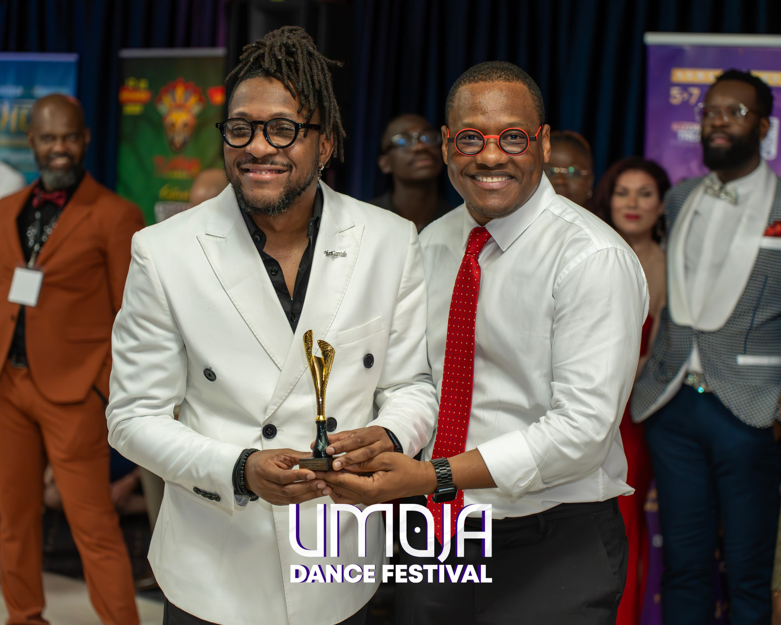 Two men smiling and holding a gold award, surrounded by other formally dressed guests at an event.