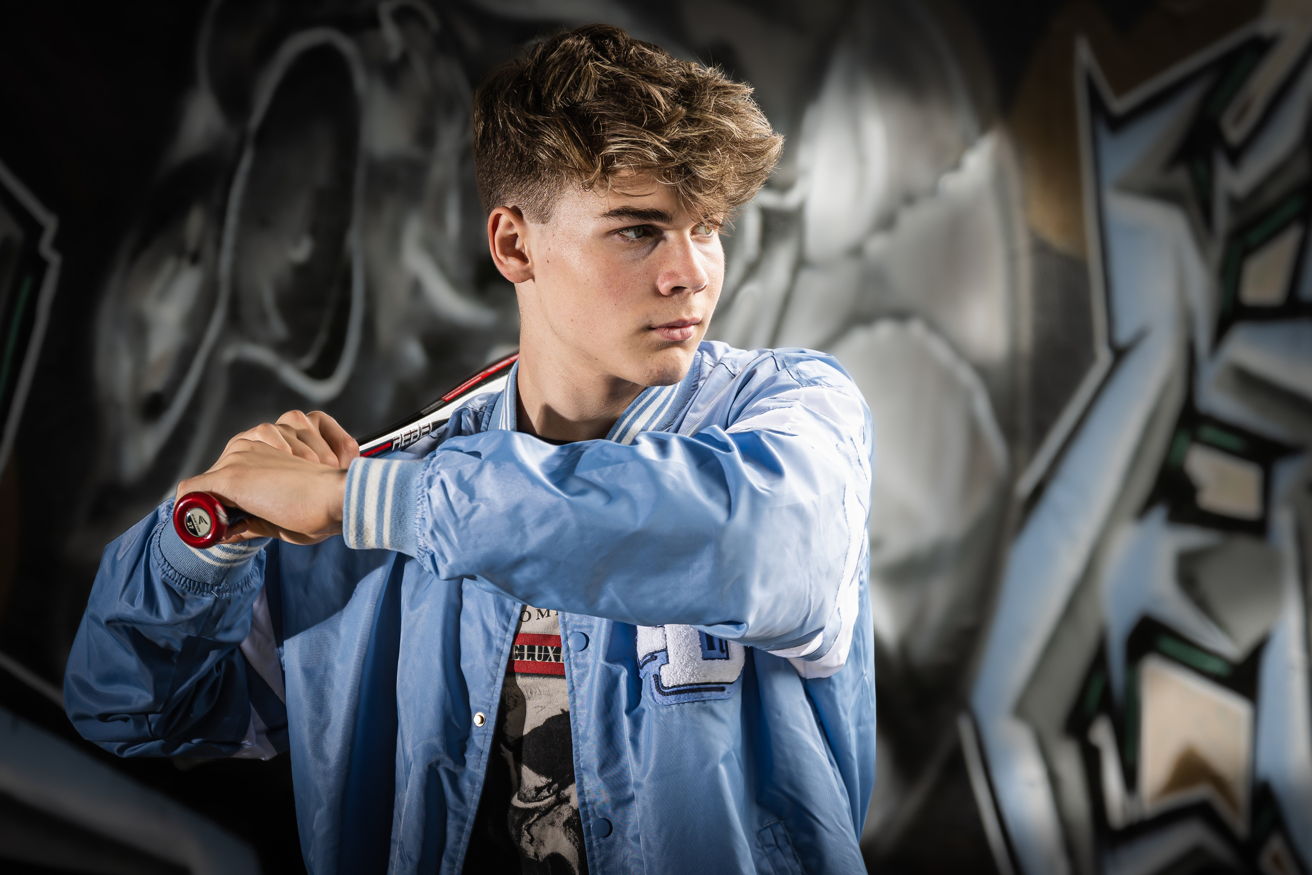 Ein Teenager in einer blauen Jeansjacke und mit einem Baseballschläger in den Händen posiert vor einer dunklen Wand mit einem Graffiti-Schädel.