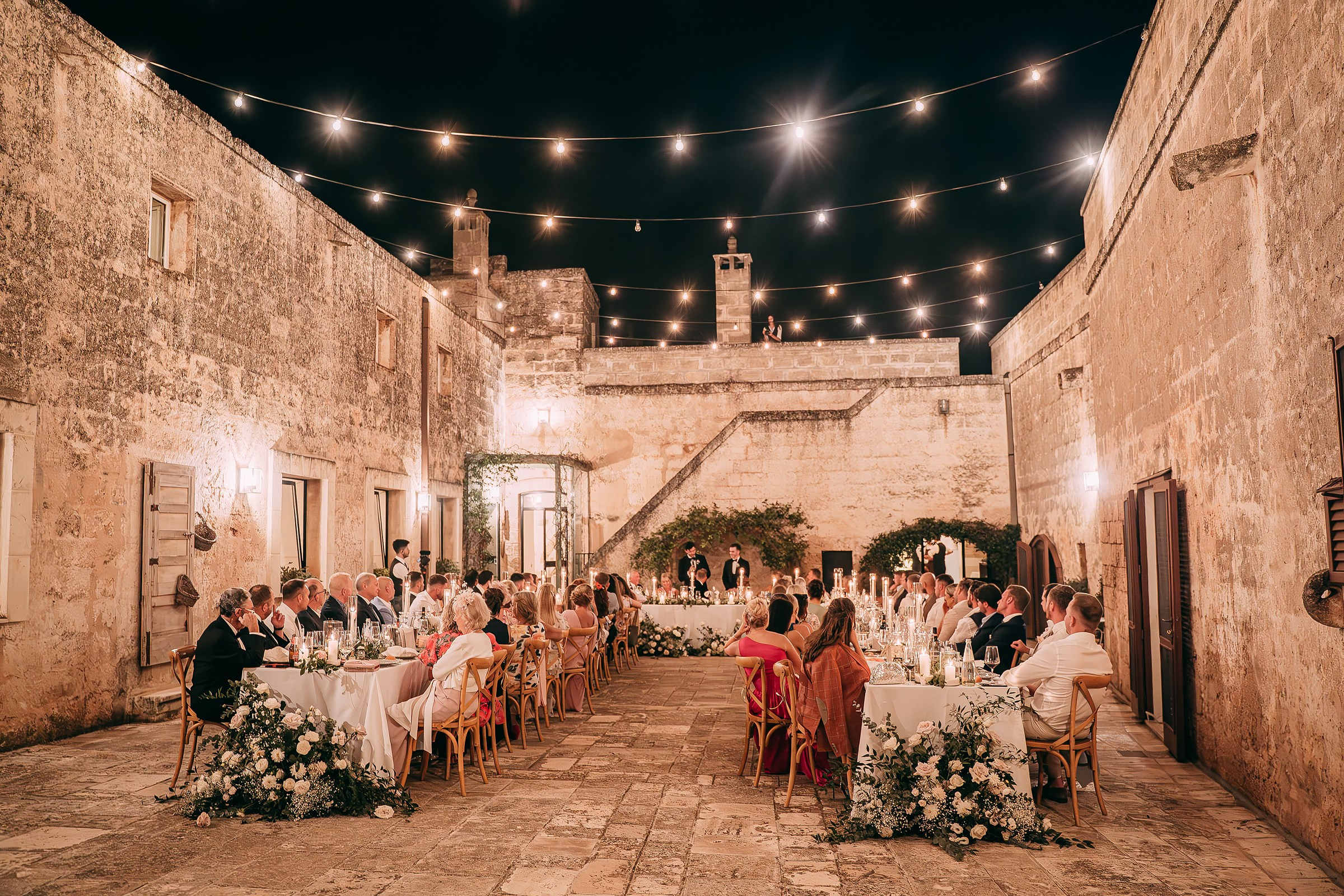 The grand evening reception setup in a historic courtyard, with long tables adorned with white flowers and candles, and guests dining under twinkling string lights against the night sky.