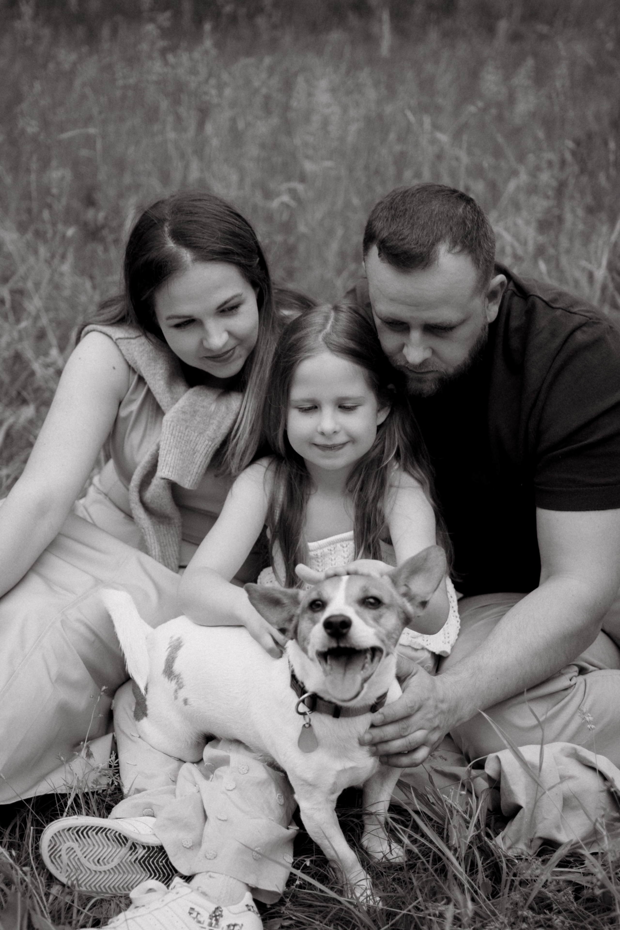 Natürliches Schwarz-Weiß-Familienfoto von Eltern und Tochter mit Hund im Grünen.