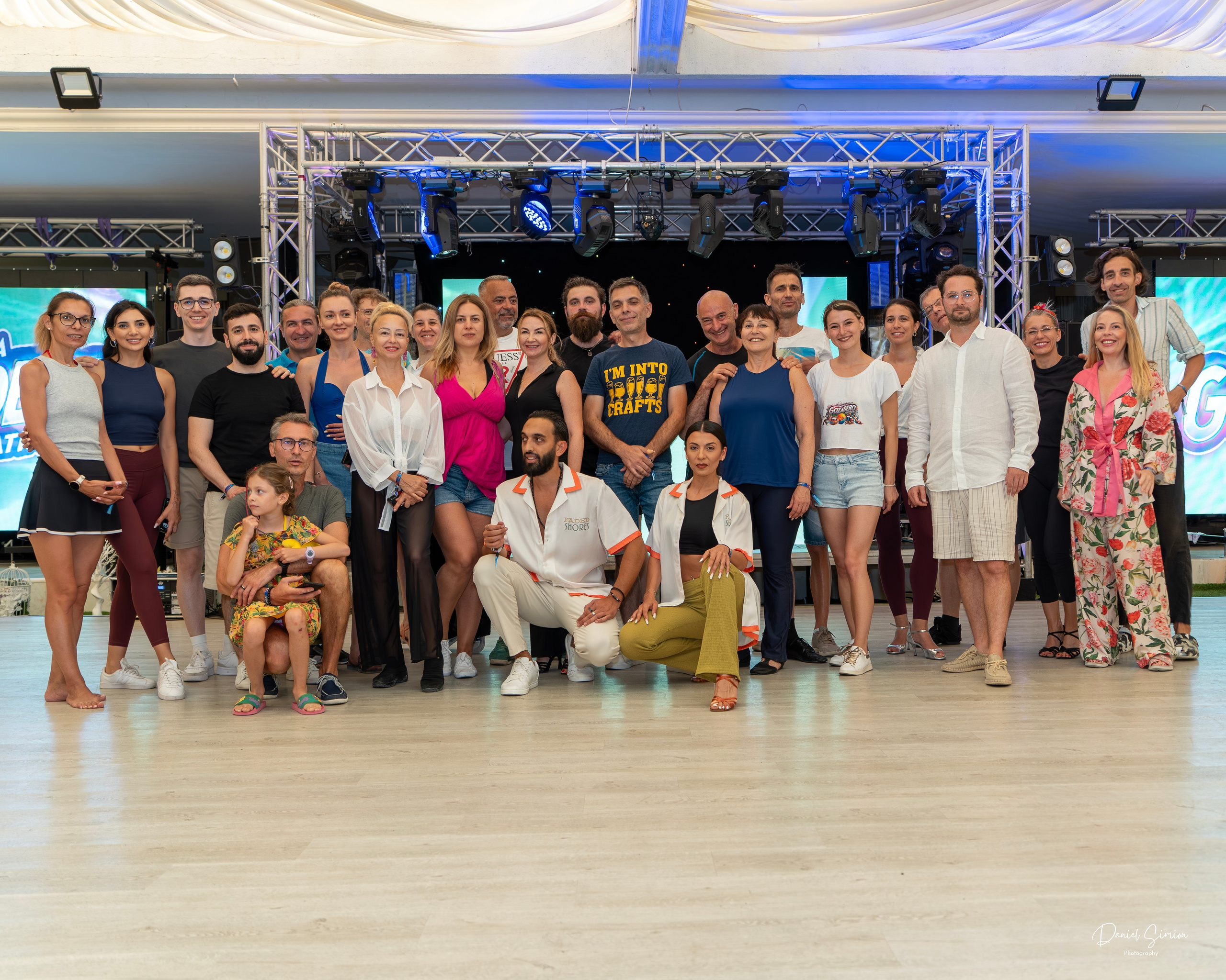 Large group of adults and one child posing in a well-lit indoor space with stage equipment.