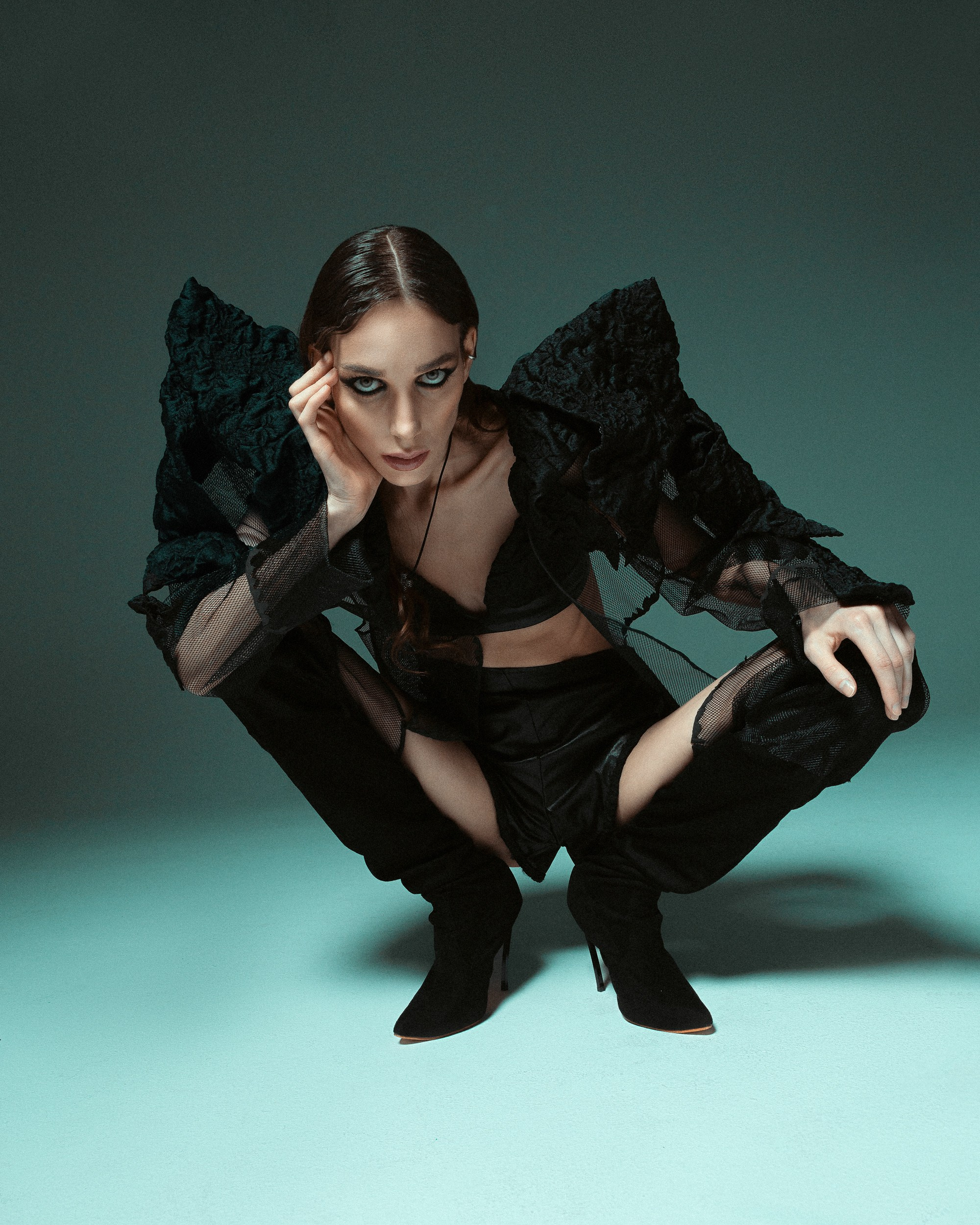 Fashion model wearing dramatic black clothing, posing in a crouched position on a teal studio floor