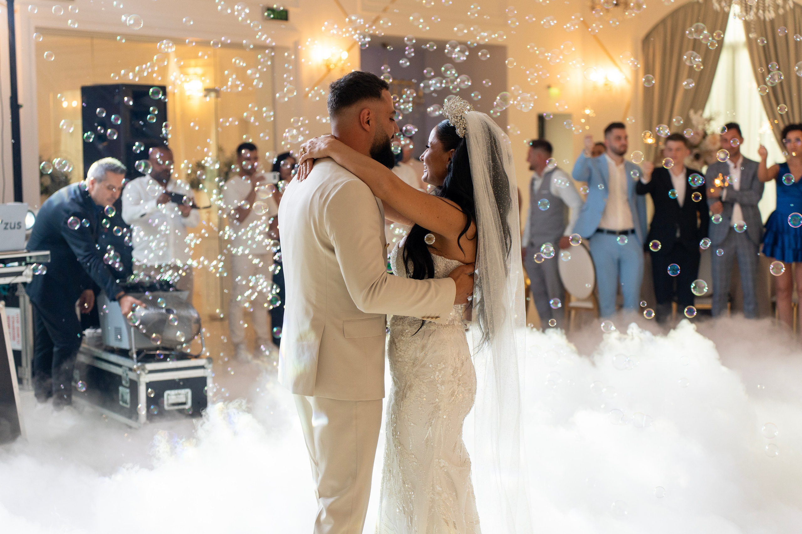 Bride and groom dancing surrounded by bubbles and fog at wedding reception with guests in background.