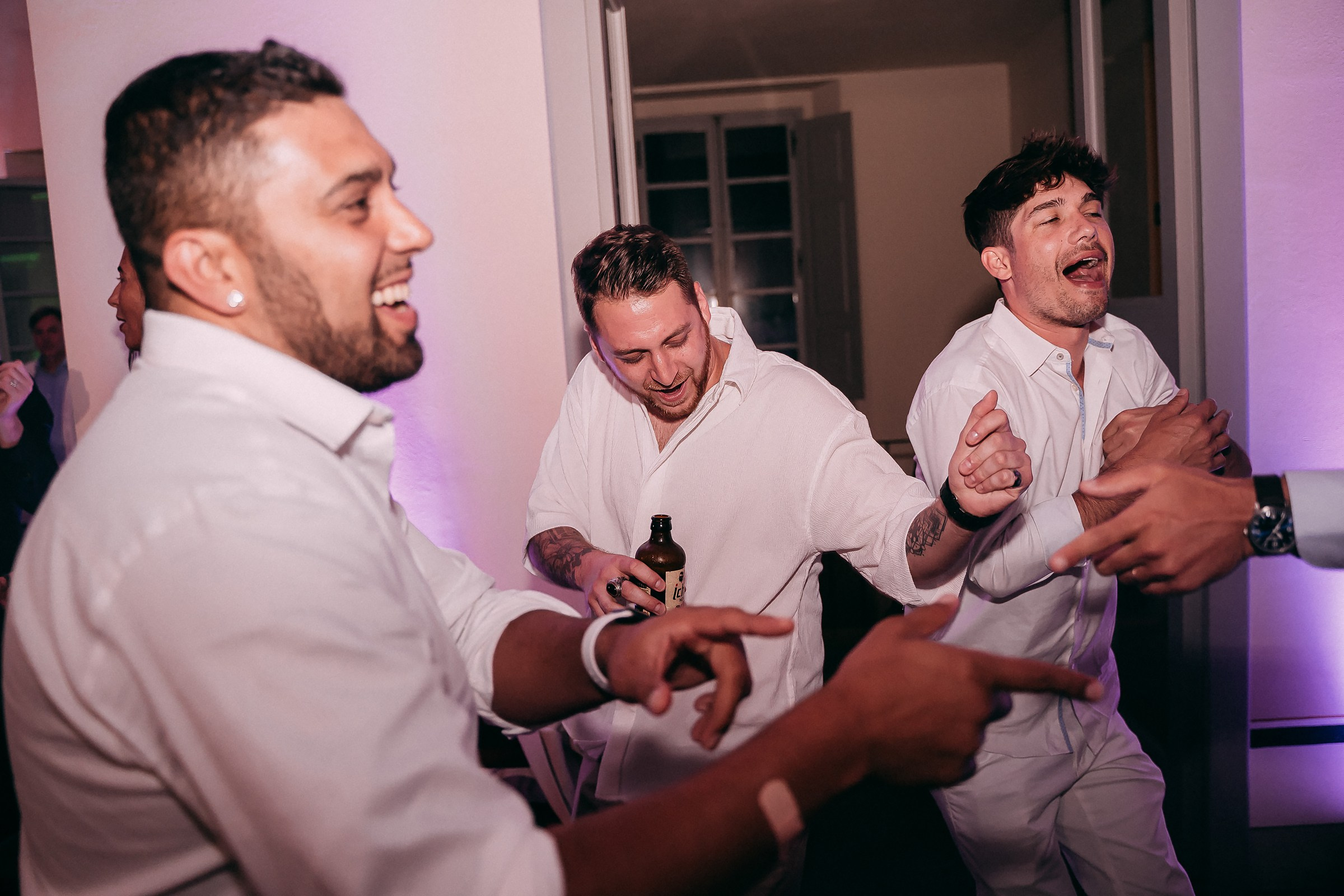 A lively group of groomsmen laugh and dance together, showcasing their camaraderie and joy in the vibrant atmosphere of the wedding party.