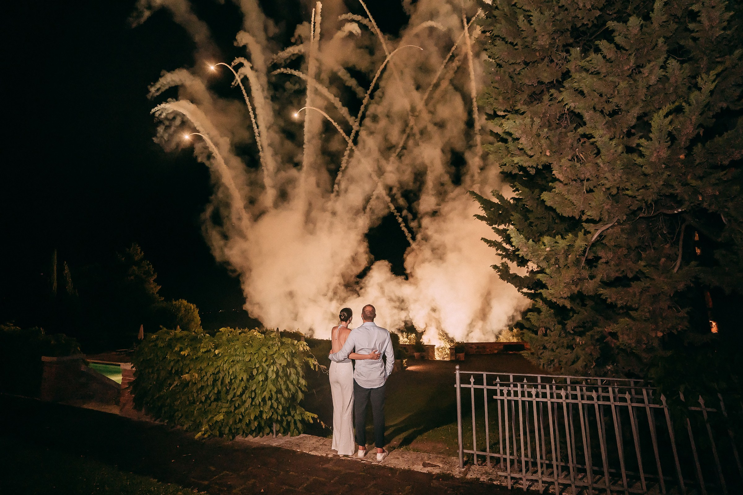 A couple standing arm-in-arm under the night sky, watching an enchanting display of golden fireworks illuminating the event.