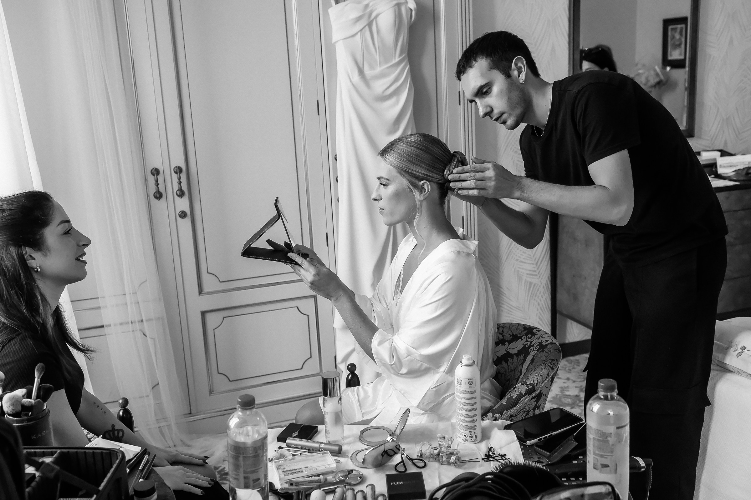 A bride in a robe surrounded by her bridal team, with a stylist adjusting her hair and a bridesmaid holding her shoes; the room is filled with wedding preparation energy.