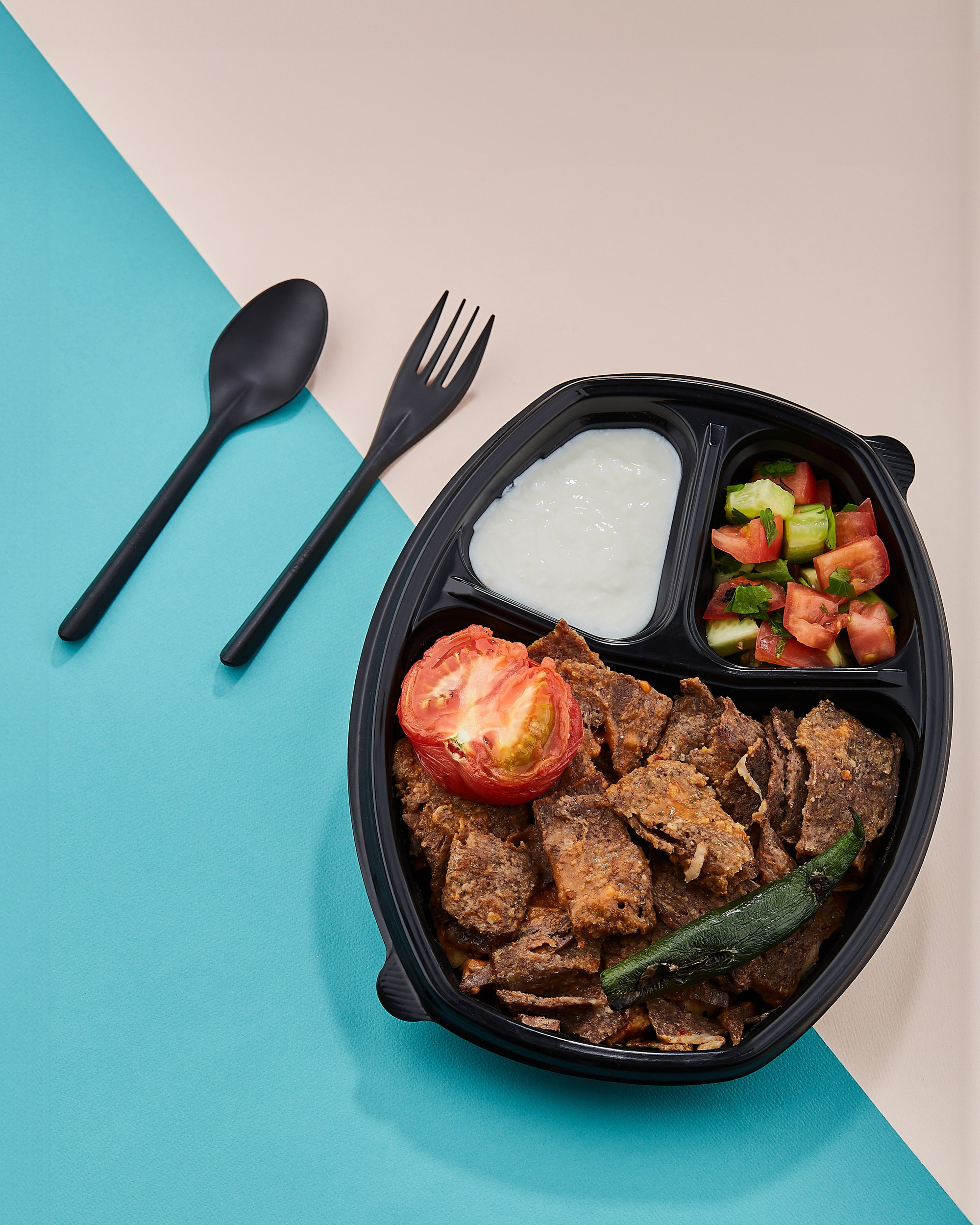 Açıq mavi və açıq çəhrayı rənglərlə diaqonal olaraq iki hissəyə bölünmüş fonda tək bir qara plastik qabda yemək görünür. Qabda doğranmış ət, qızardılmış pomidor, yaşıl bibər, bir az salat və ağ qatıq və ya sous var. Qabın yanında qara plastik qaşıq və çəngəl var.