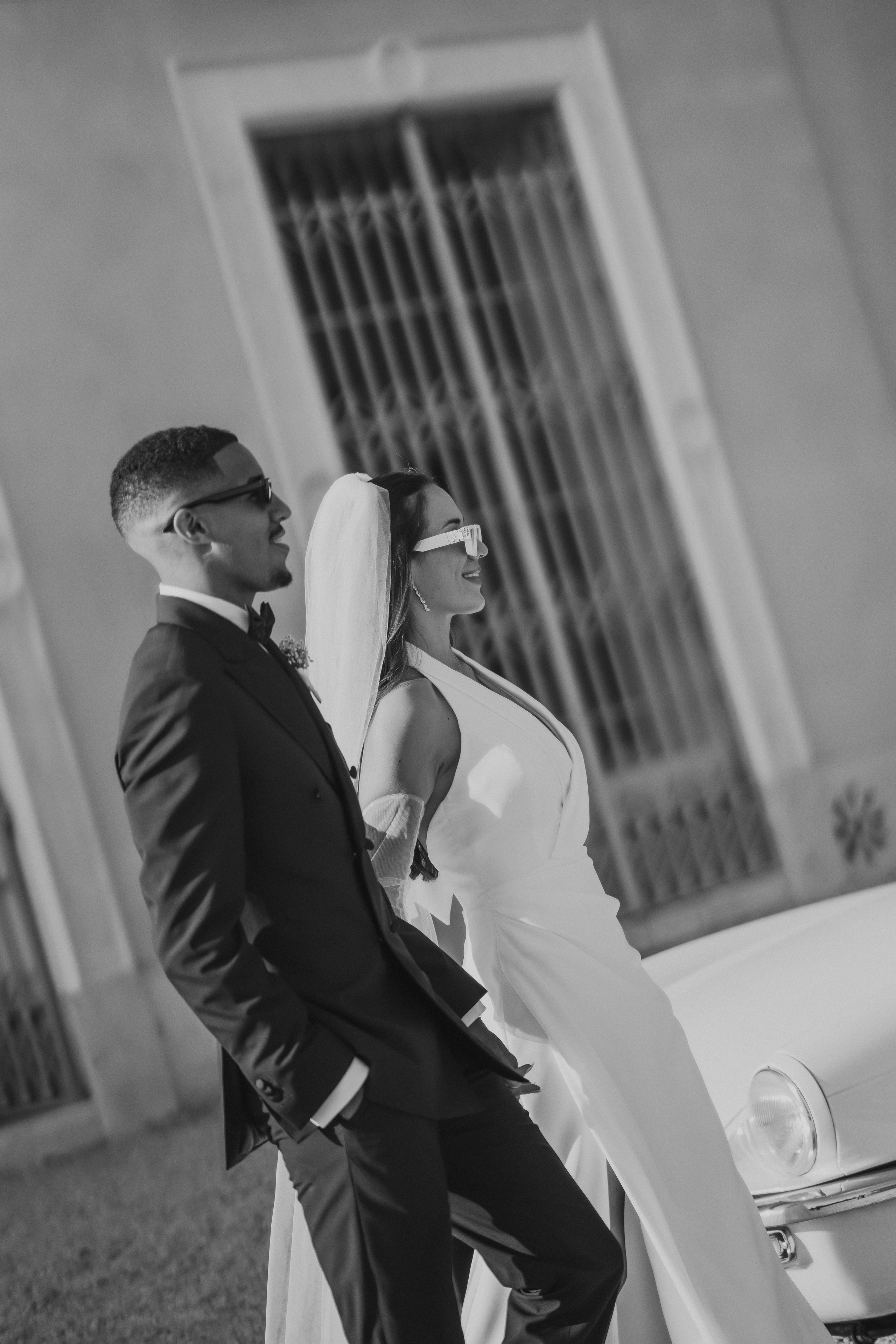 Casal em pose editorial, com vestido moderno e óculos de sol, frente a palácio em Itália.