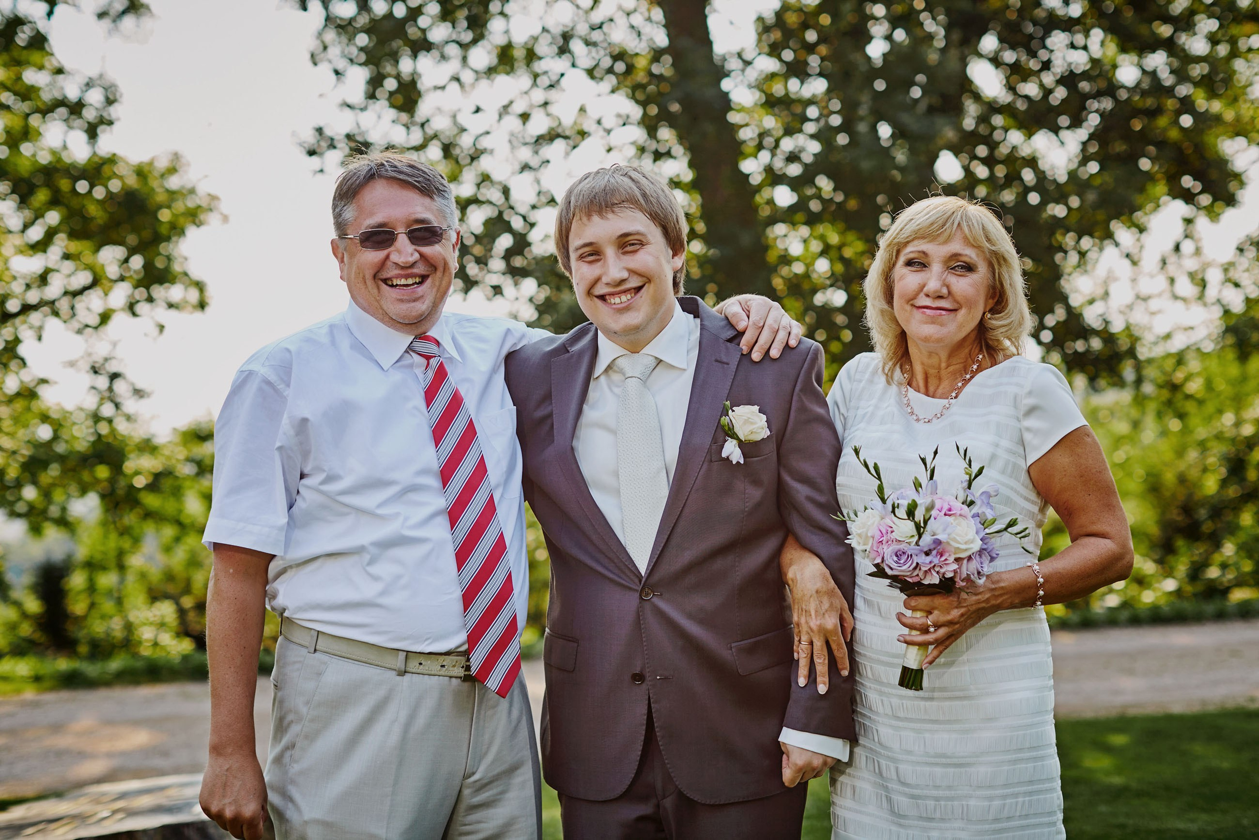The groom's parents cuddle up with him as they pose for a photo following his summer, outdoor wedding on the grounds of the Chateau Mcely.
