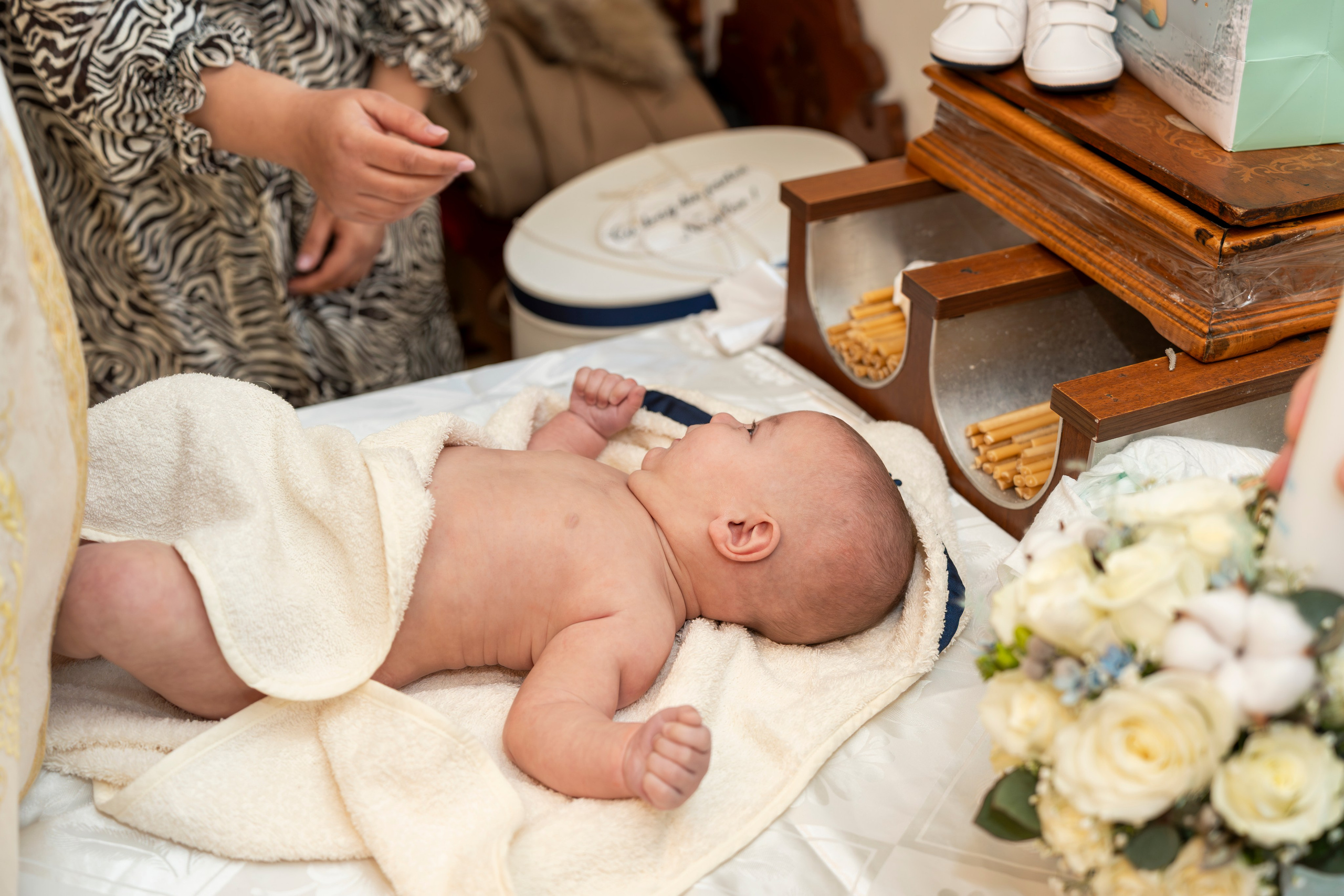 Woman comforting a crying baby wearing a floral dress indoors with blurred people in background.