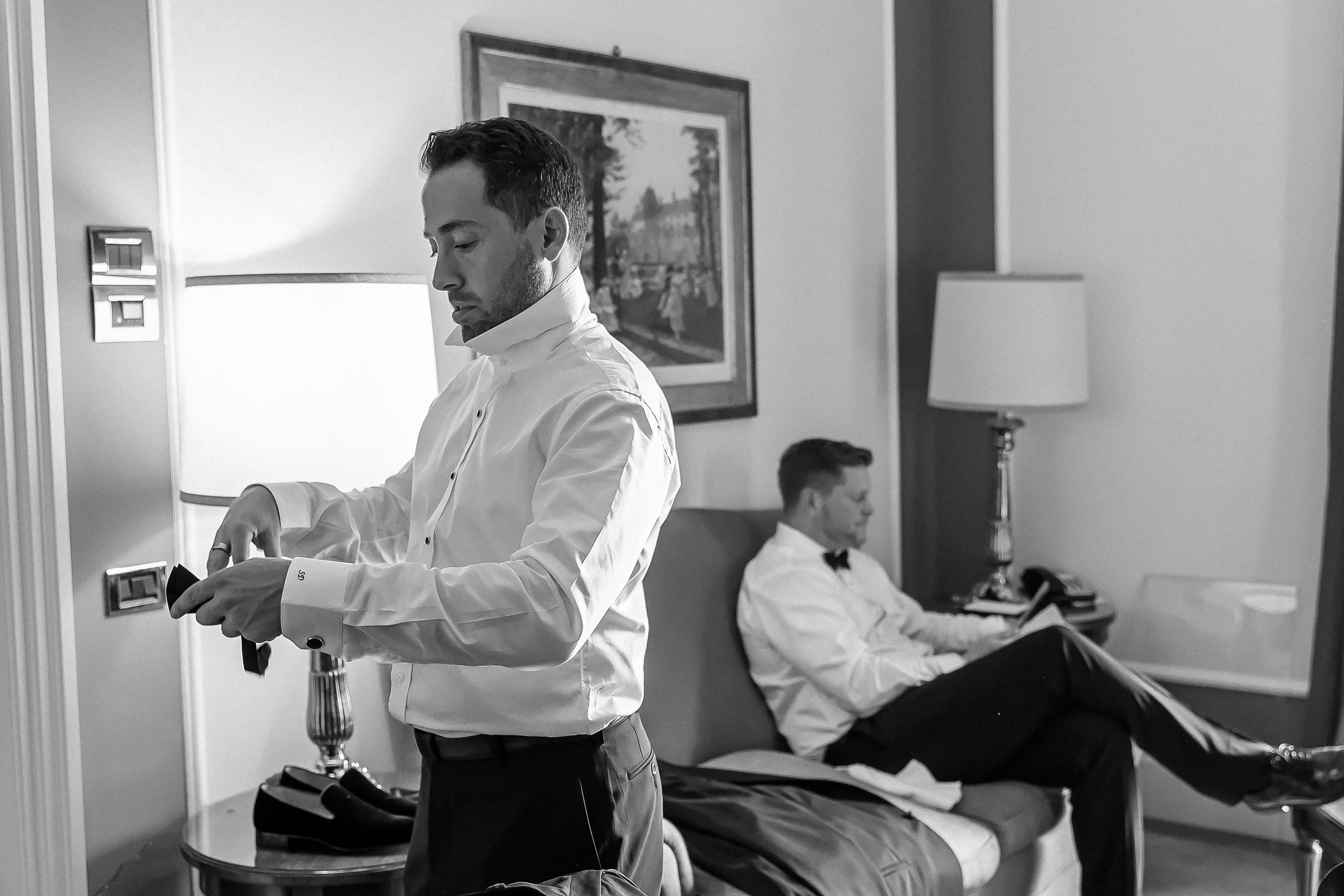 A man in white shirt getting ready while other man sits on the sofa in luxury room.