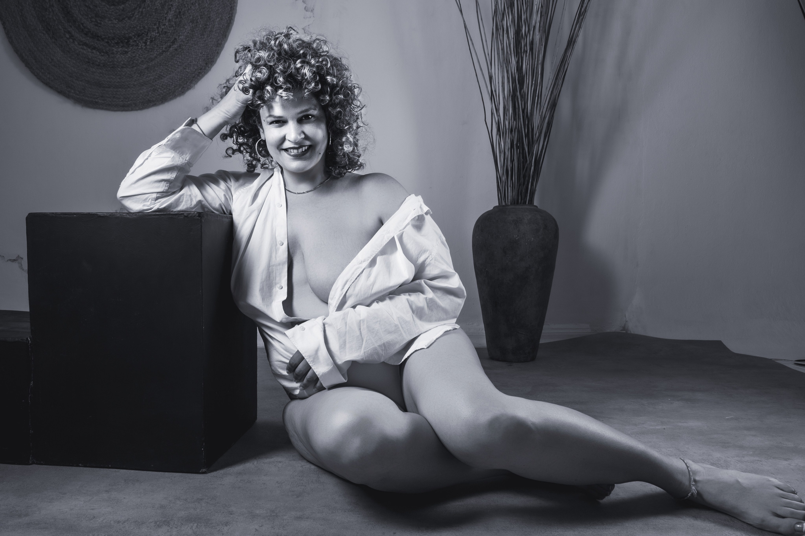 Mujer de pelo rizado sentada en el suelo con camisa blanca abierta junto a un jarrón, boudoir en blanco y negro