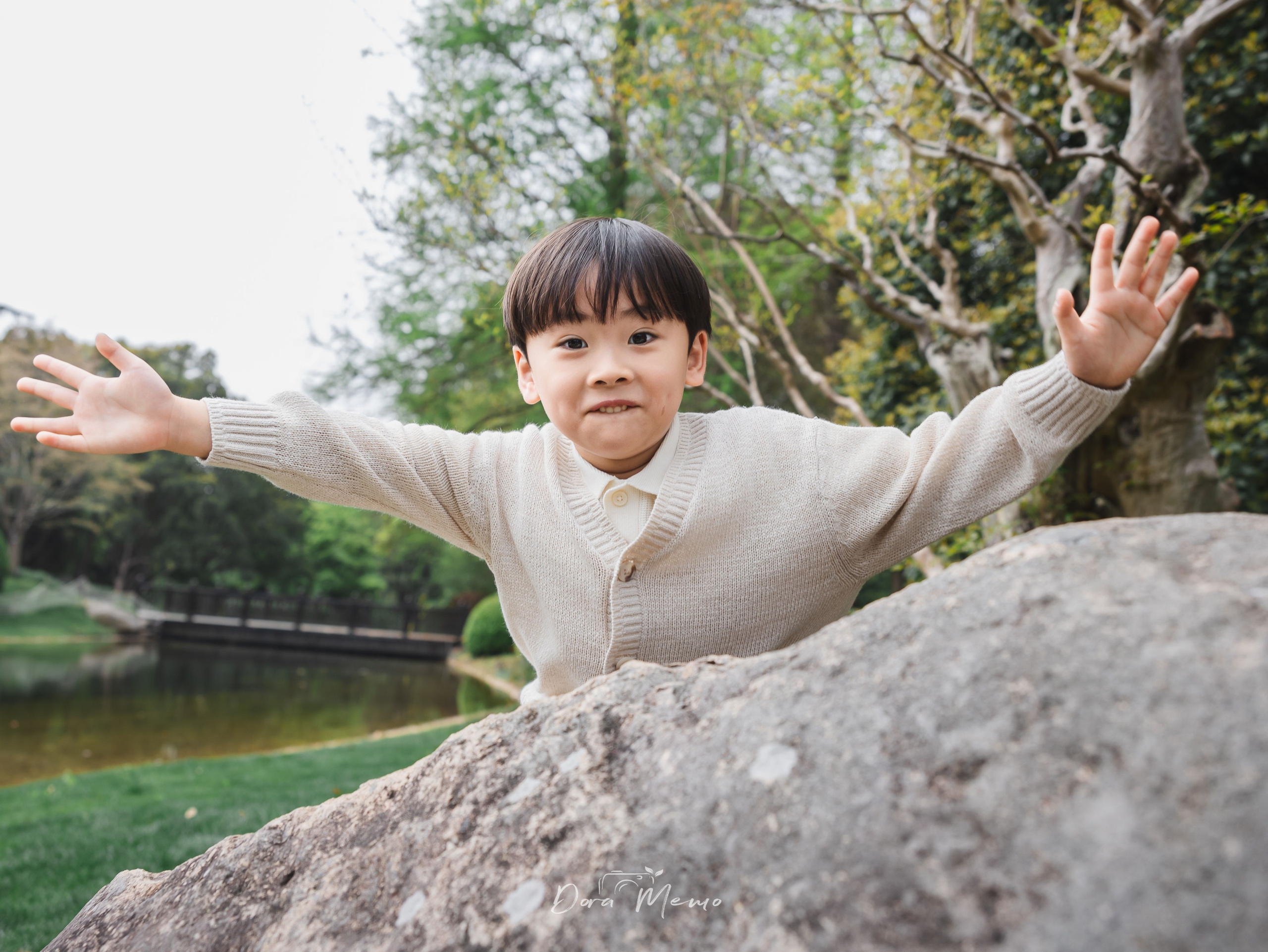A boy stands behind a rock with arms spread wide, beaming at the camera