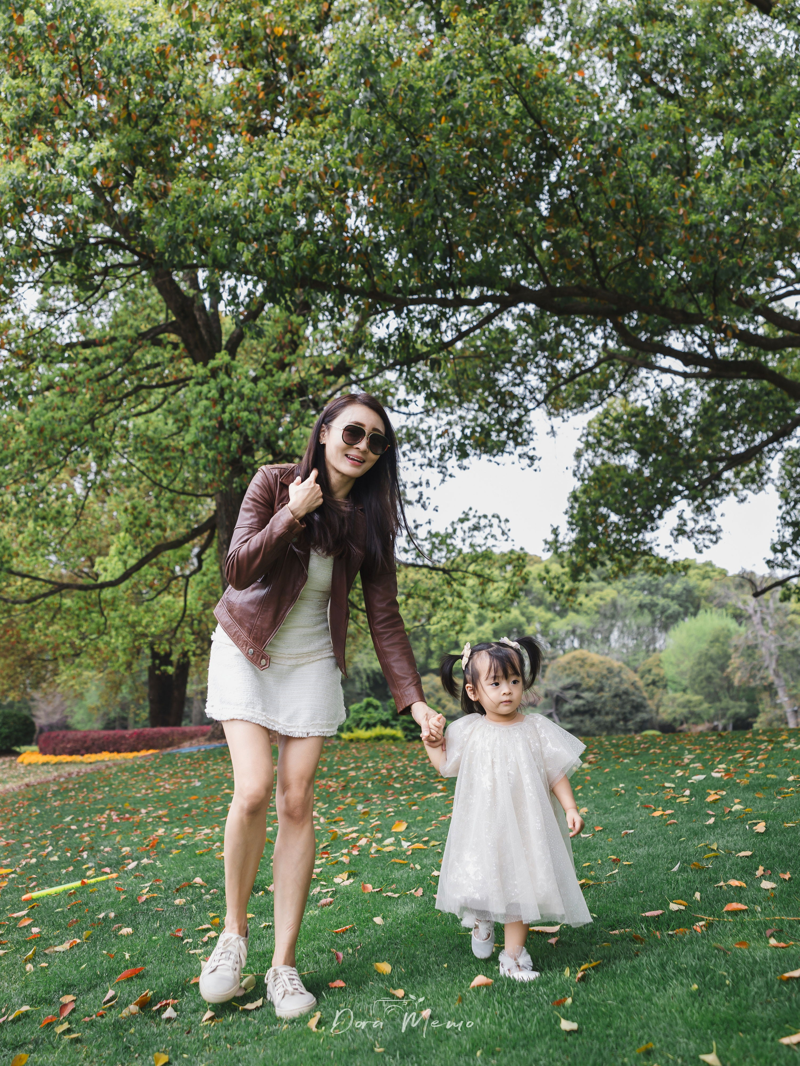 A mother holds her toddler daughter's hand as they walk through fallen leaves