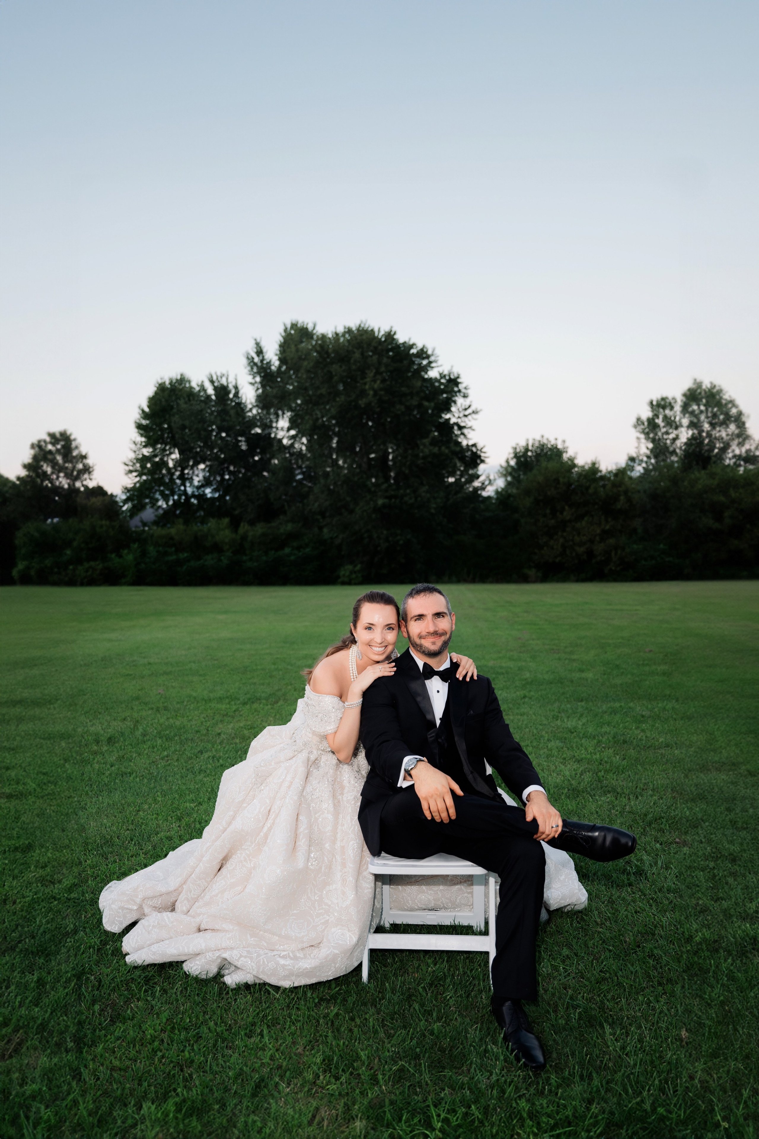 a bride and groom sitting on a white bench in a field