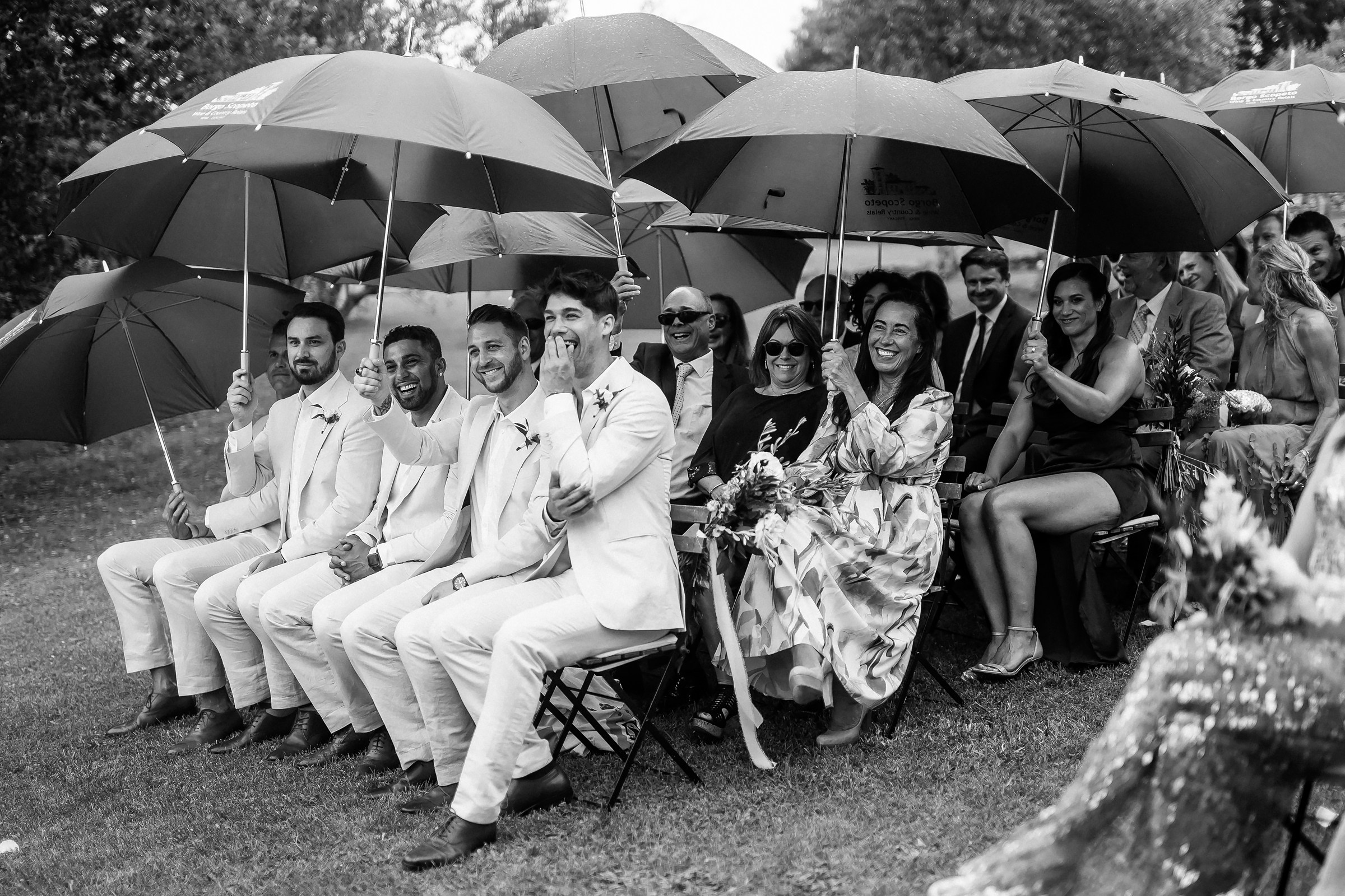 A candid black-and-white capture of the groomsmen and guests under umbrellas, sharing laughter during the ceremony.