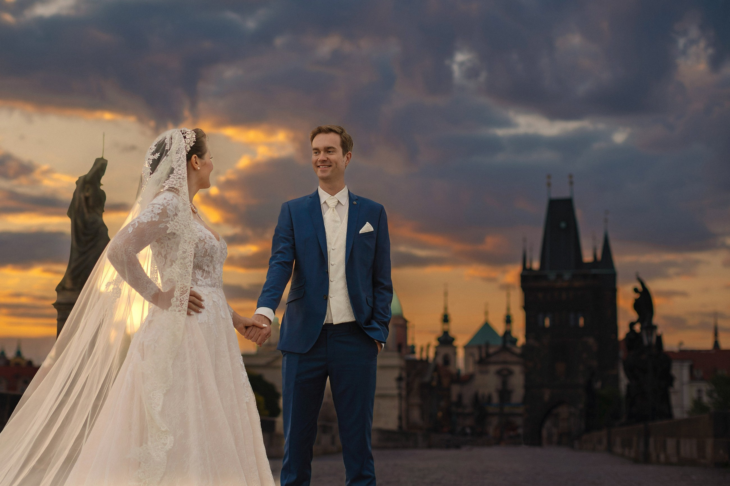 A bride and groom lovingly look towards each other as they stand underneath gas lamps as the sky flares vividly behing them during their Charles Bridge wedding photo session at dawn.