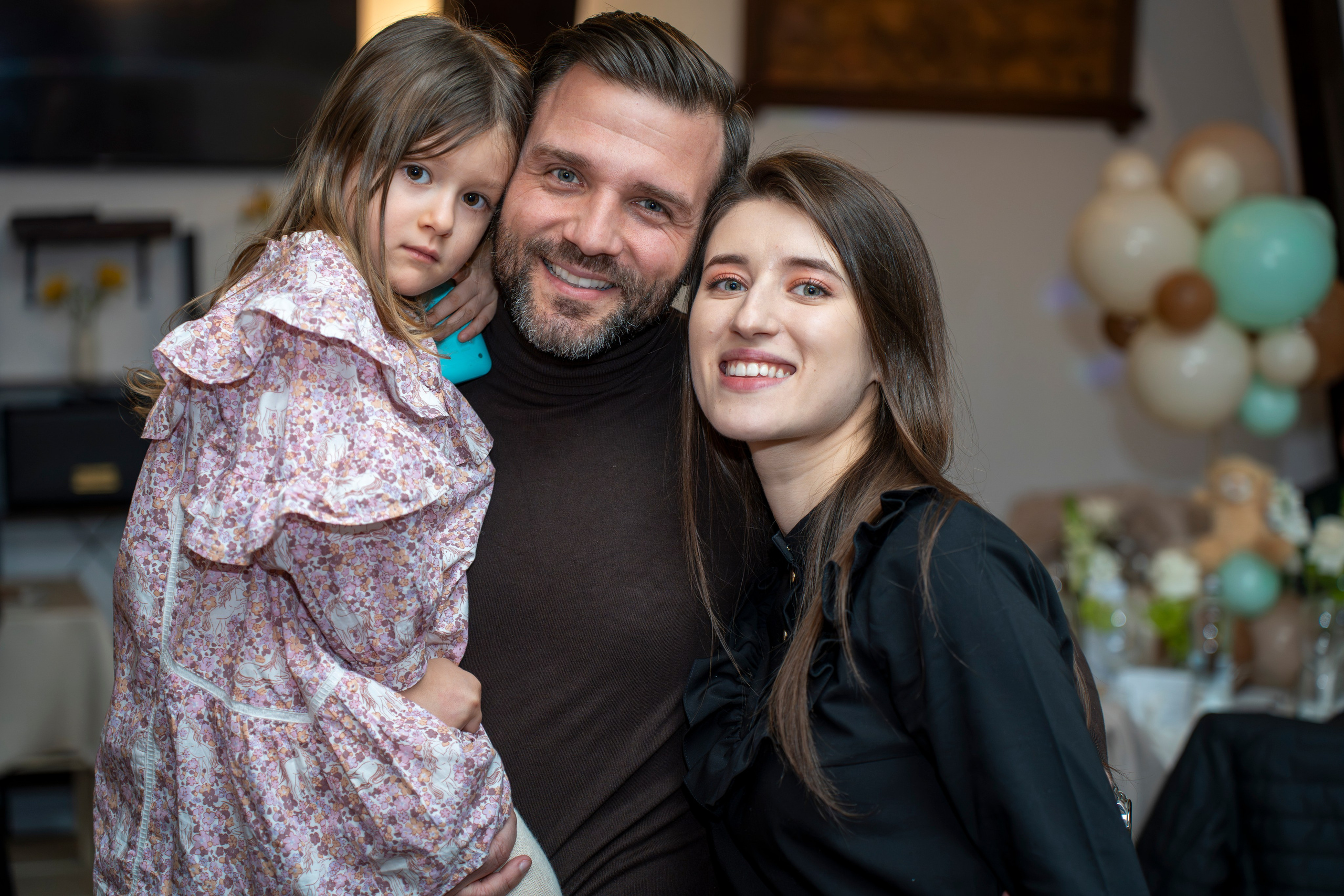 Man and woman smiling next to a young girl with floral dress indoors with balloon decorations.