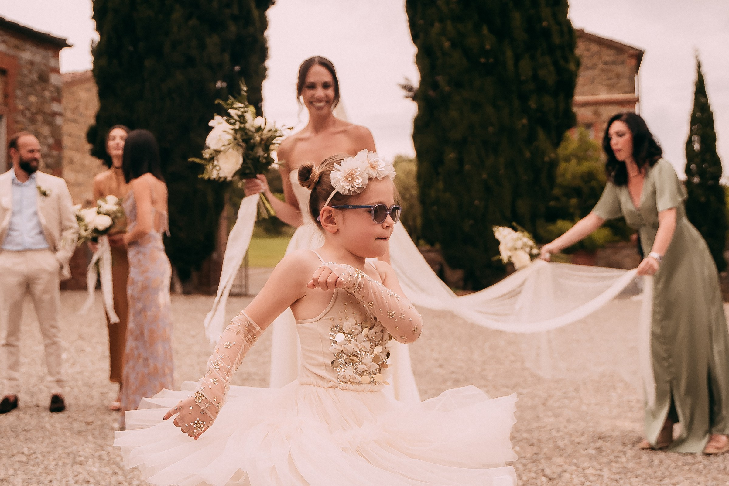 A flower girl in a sparkly cream dress and sunglasses spins joyfully as the bride smiles in the background.