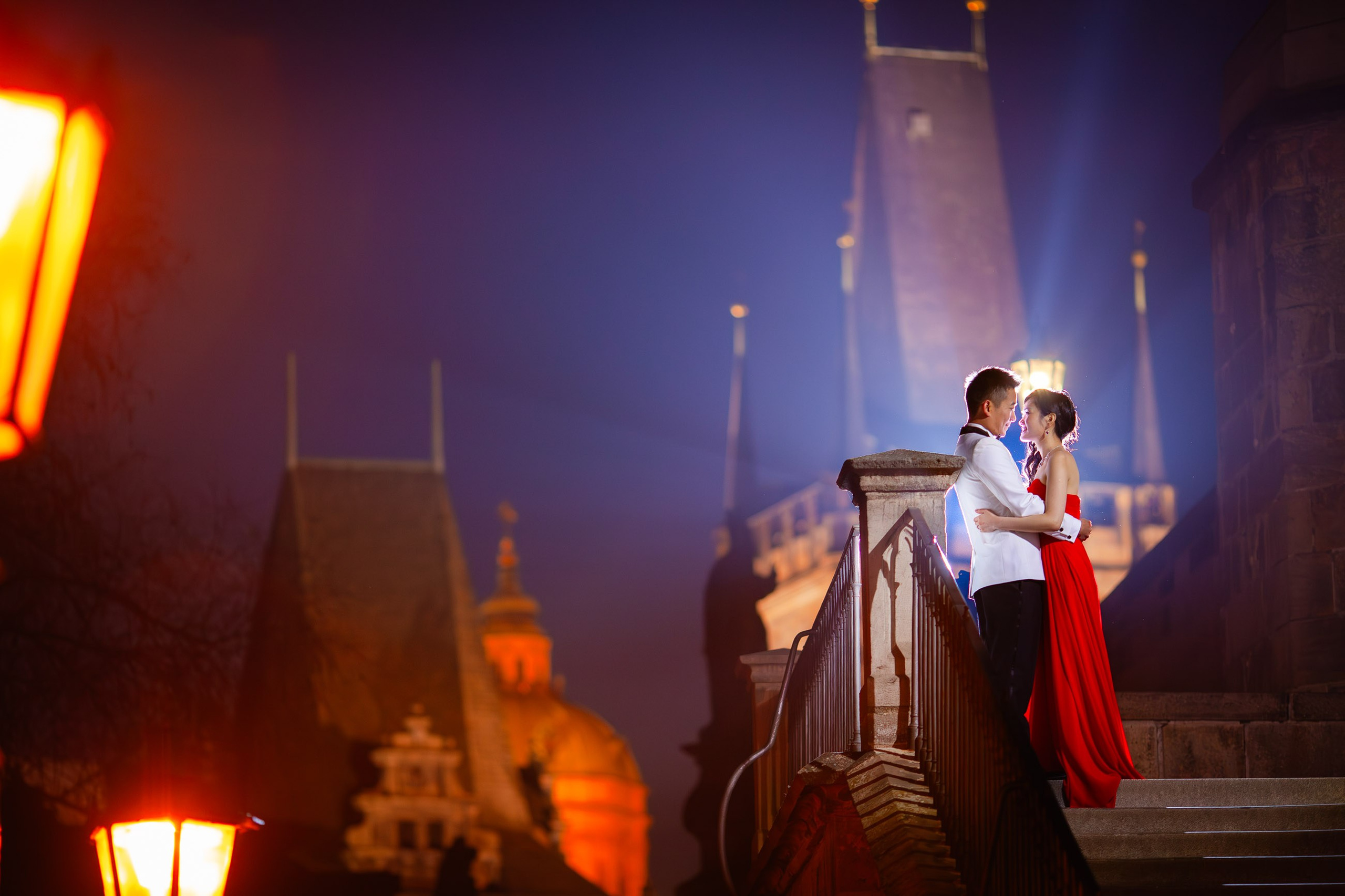 A smiling Hong Kong woman wearing a stylish red evening dress and her white tuxedo wearing man stand atop the Kampa steps as they are illuminated by the gas lamps of the Charles Bridge.