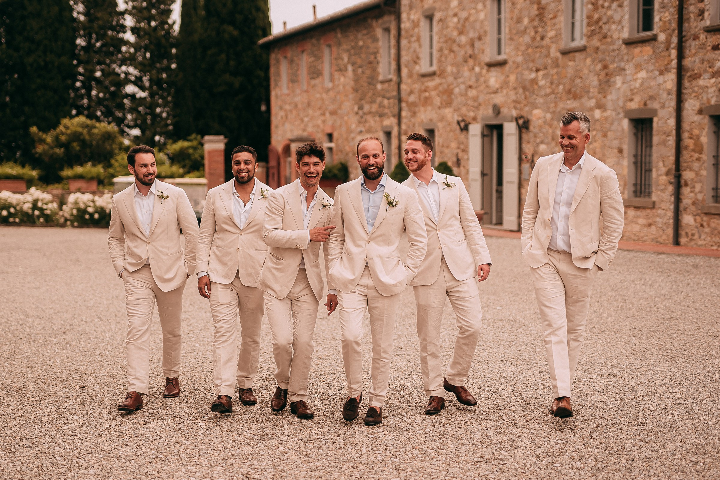 Groomsmen in light suits pose formally in front of a rustic stone building, radiating confidence and charm.