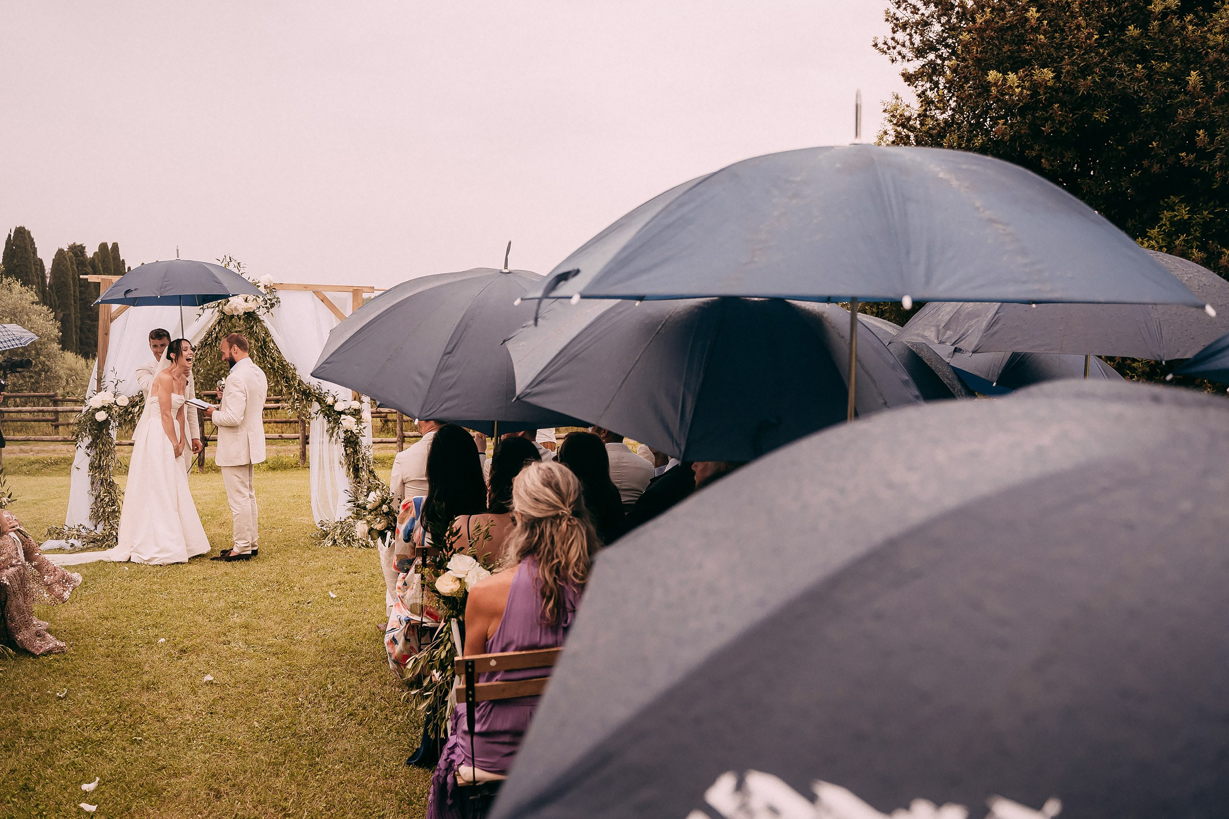 Wedding ceremony continues under umbrellas, creating an intimate atmosphere amid soft raindrops in the Tuscan countryside.