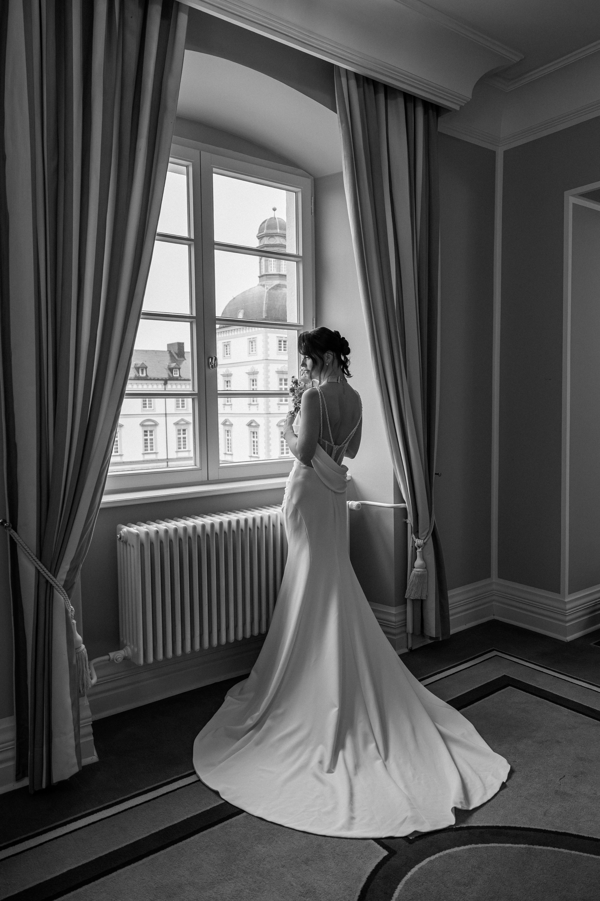 Braut im langen Kleid am Fenster, elegantes Schwarzweiß‑Portrait.