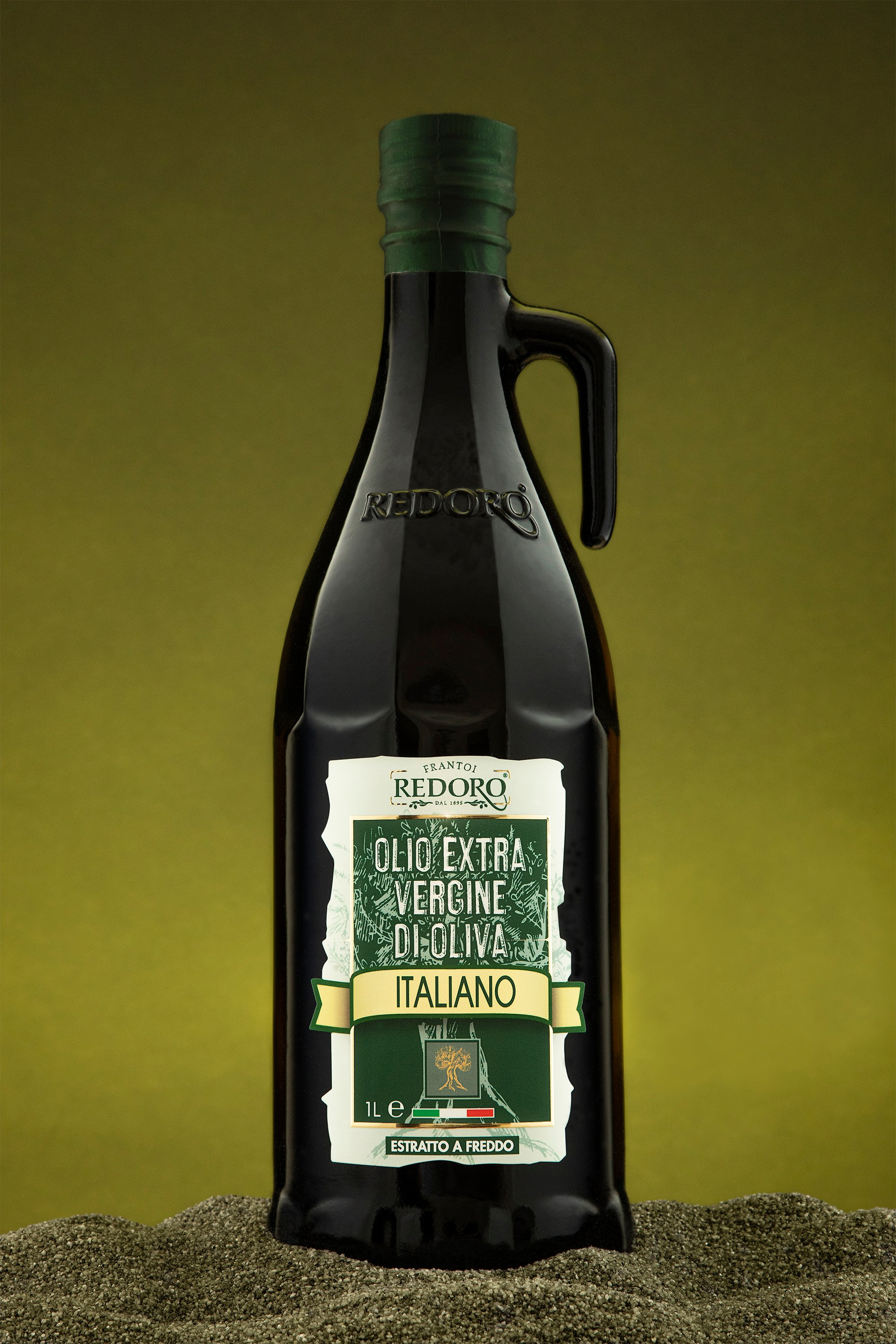 Photographer Roman Djuzev - A photo of a glass bottle of Redoro olive oil located on dark gray sand, the background is dark olive.