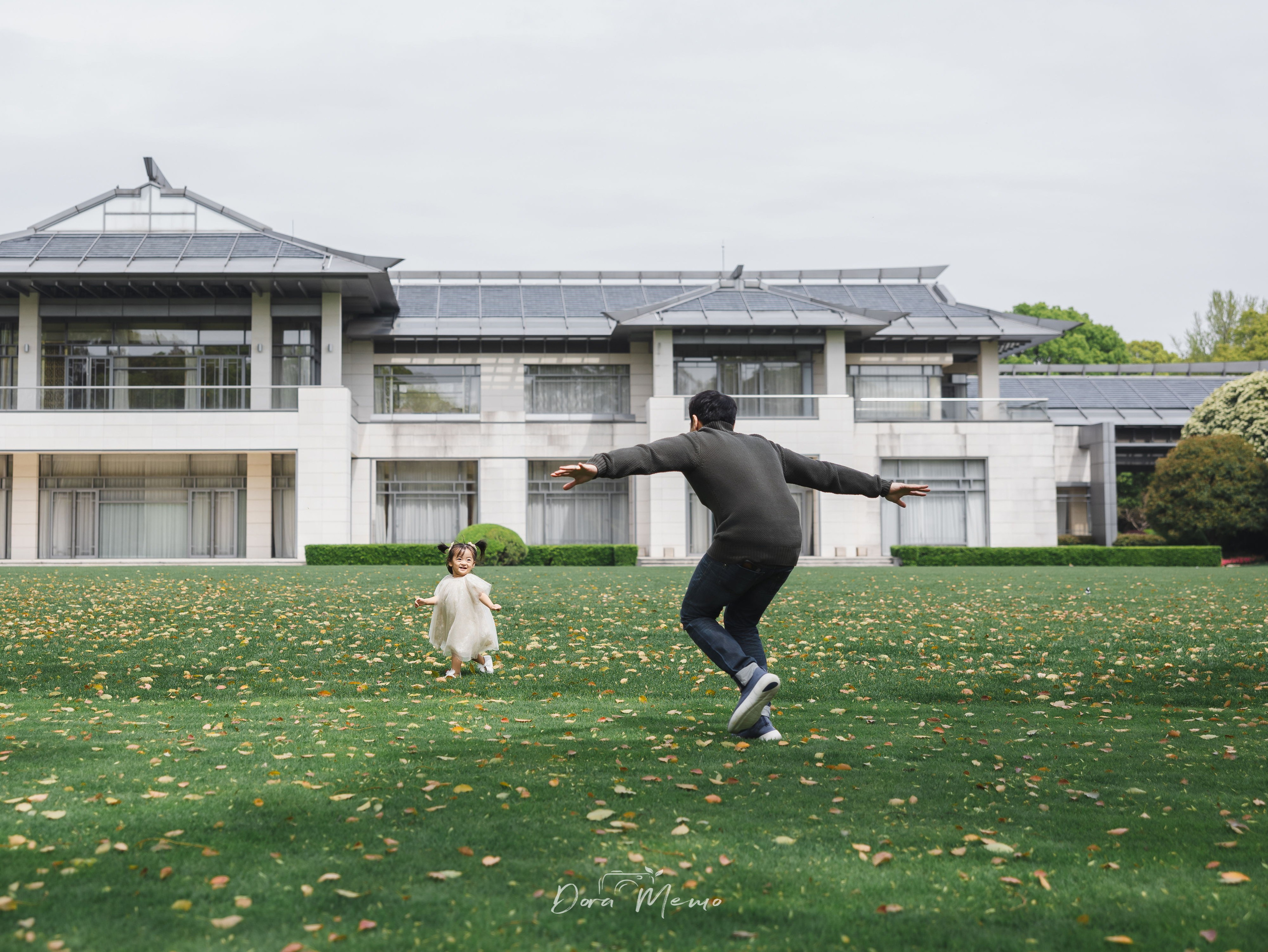 A father chases his laughing toddler daughter across a lawn in Shanghai