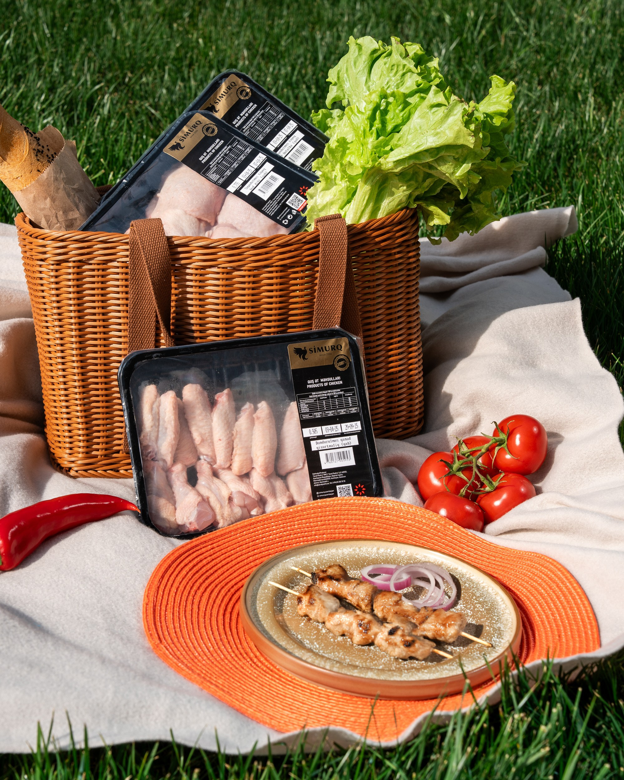 Simurq brendinə aid qablaşdırılmış toyuq qanadları piknik səbətində, yanında kabab və təzə tərəvəz – açıq hava məhsul fotoqrafiyası