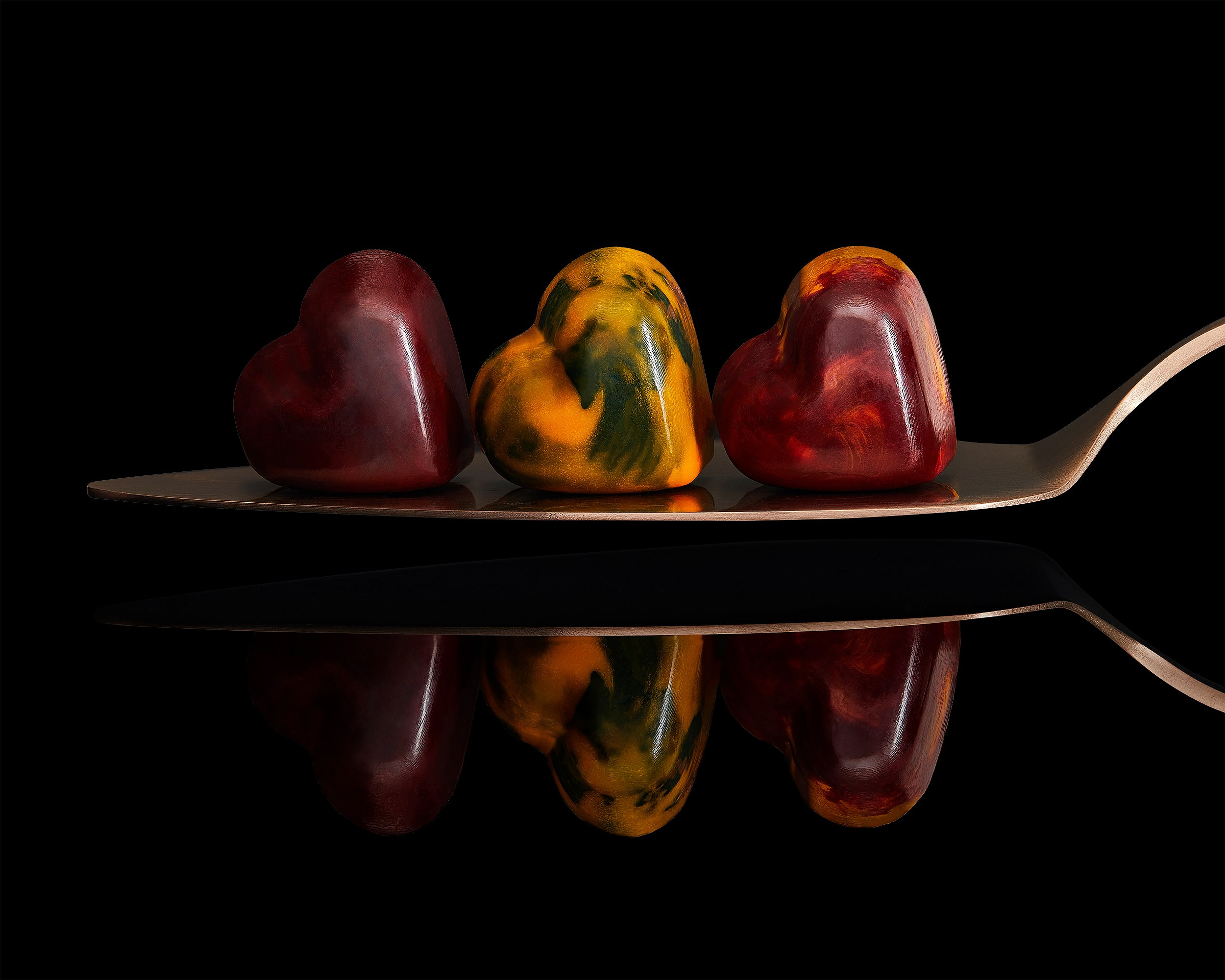 Three heart-shaped chocolate candies on a metal spoon with reflection on black background, close-up food and product photography.