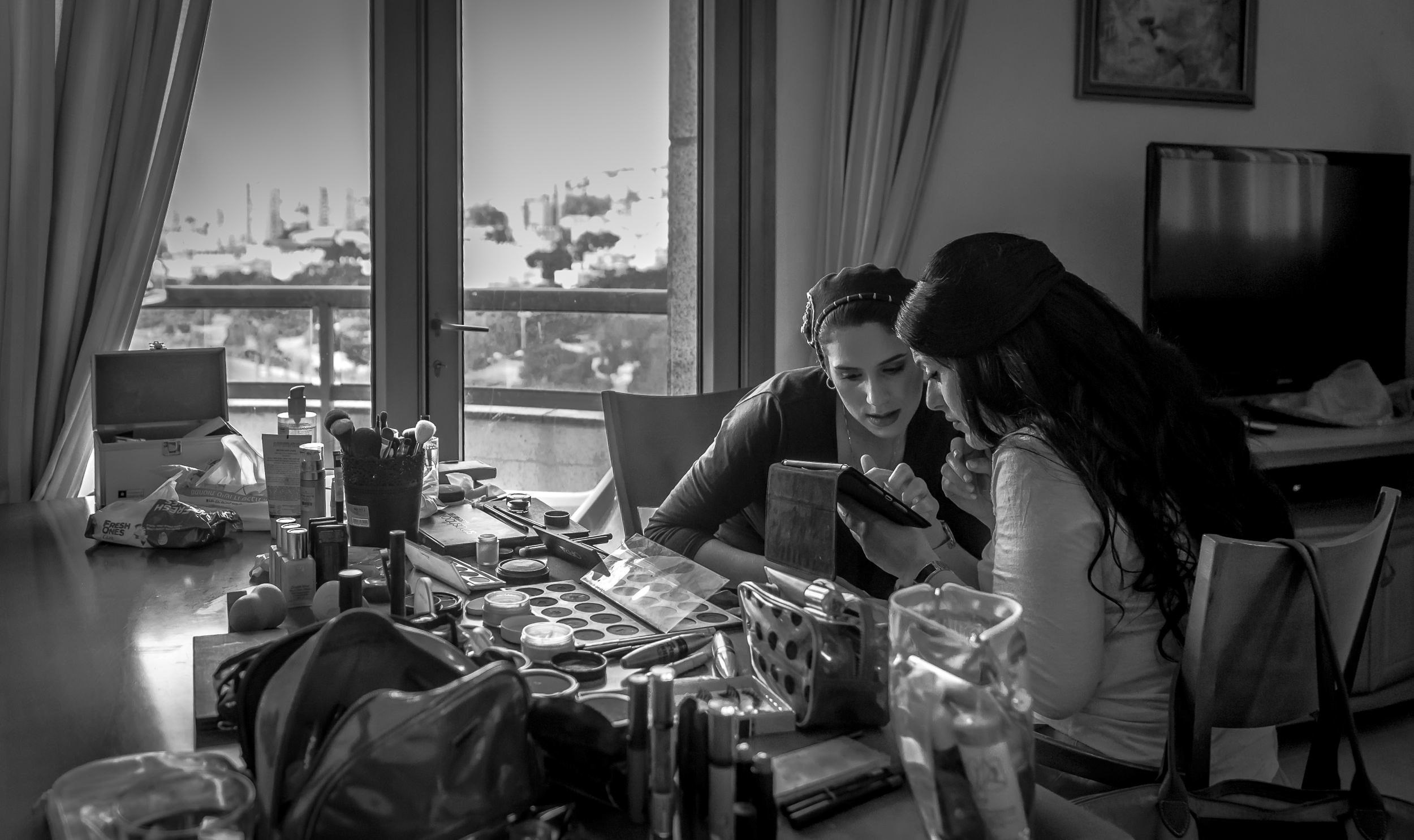 A bride and a woman look at a phone surrounded by makeup and personal items