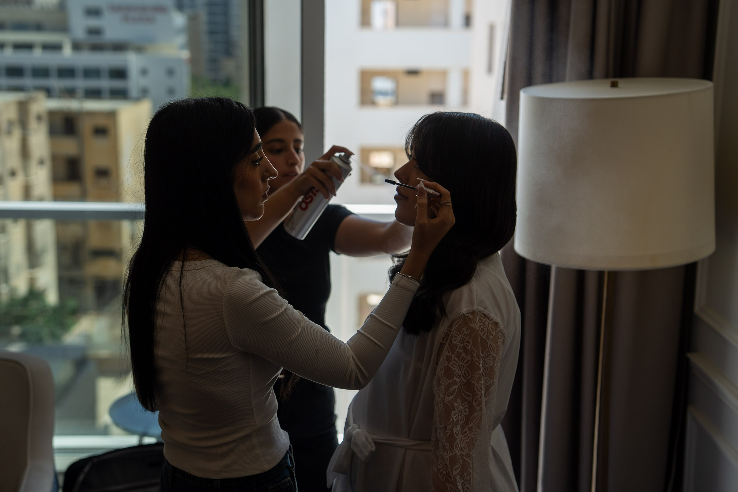 Two women help the bride with her makeup beside a window