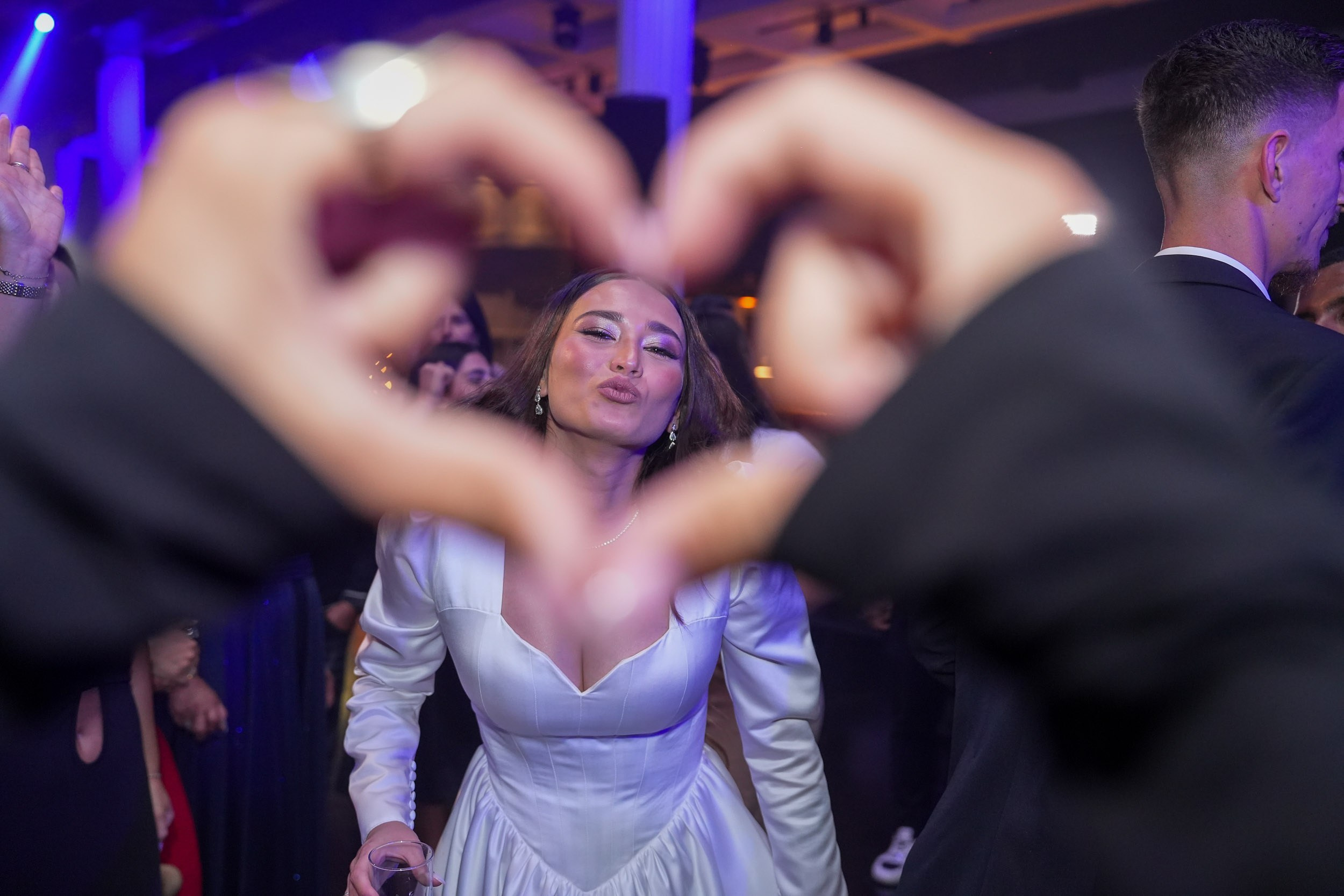 A bride smiles through a heart shape formed by guests’ hands on the dance floor