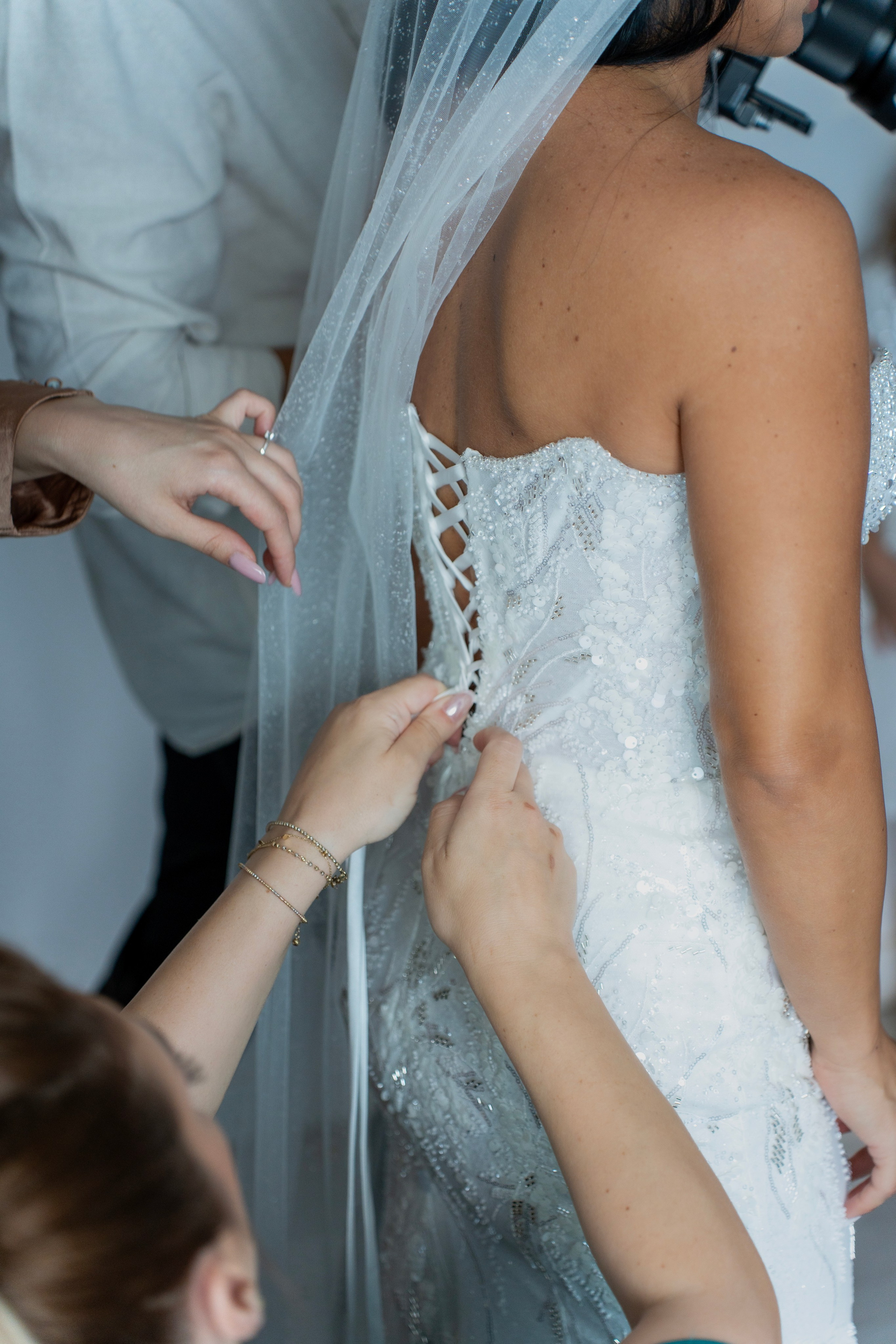 Hands adjusting laces on the back of a bride's embellished wedding dress with veil.
