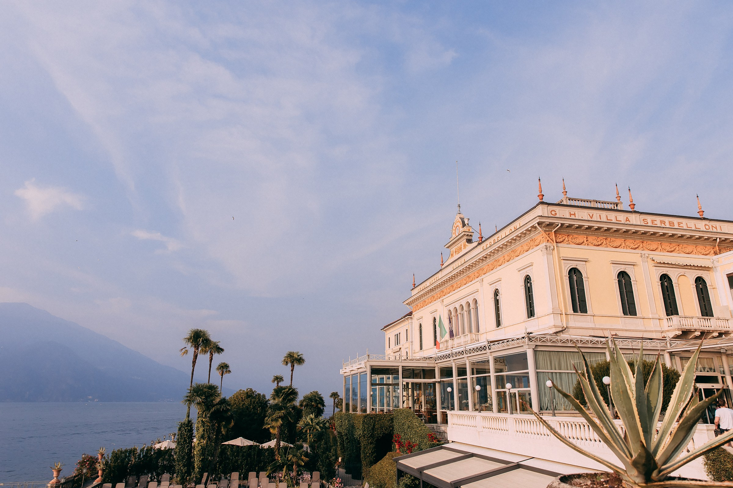 A grand view of Villa Serbelloni at Lake Como, showcasing its elegant architecture, surrounded by lush greenery and overlooking the serene waters of the lake under a clear sky.