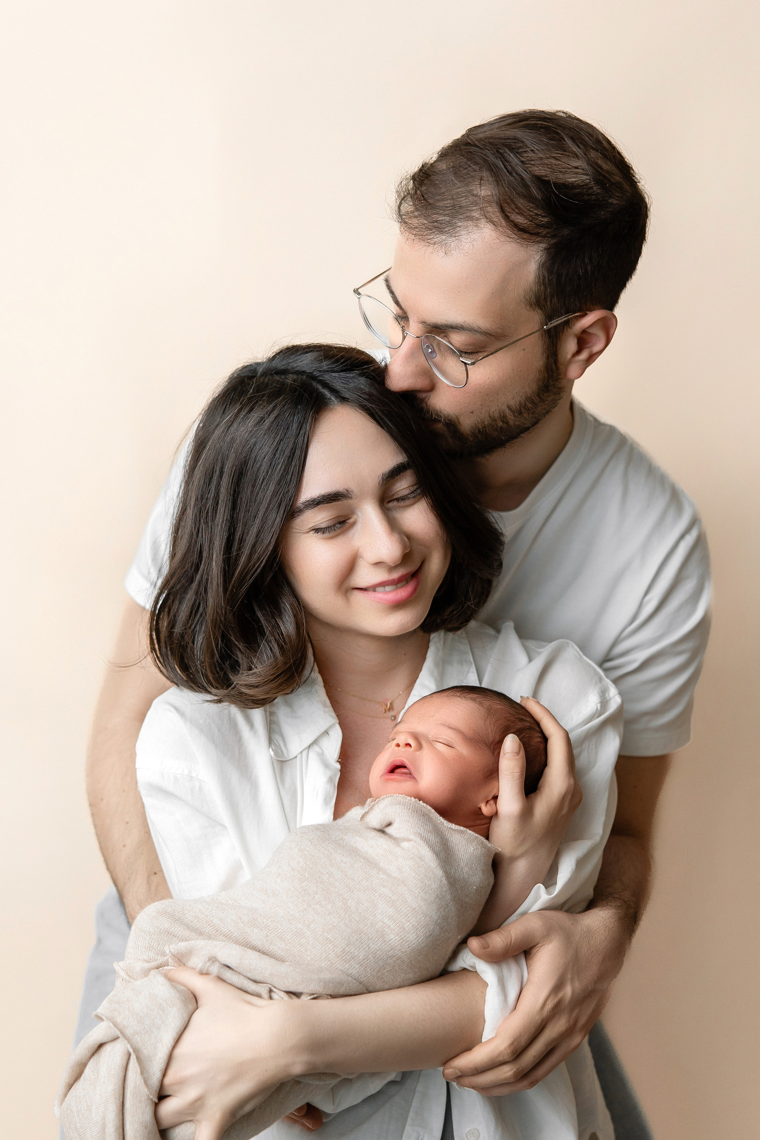 Happy young parents lovingly holding their newborn baby wrapped in a beige swaddle — intimate family portrait in a soft studio setting.