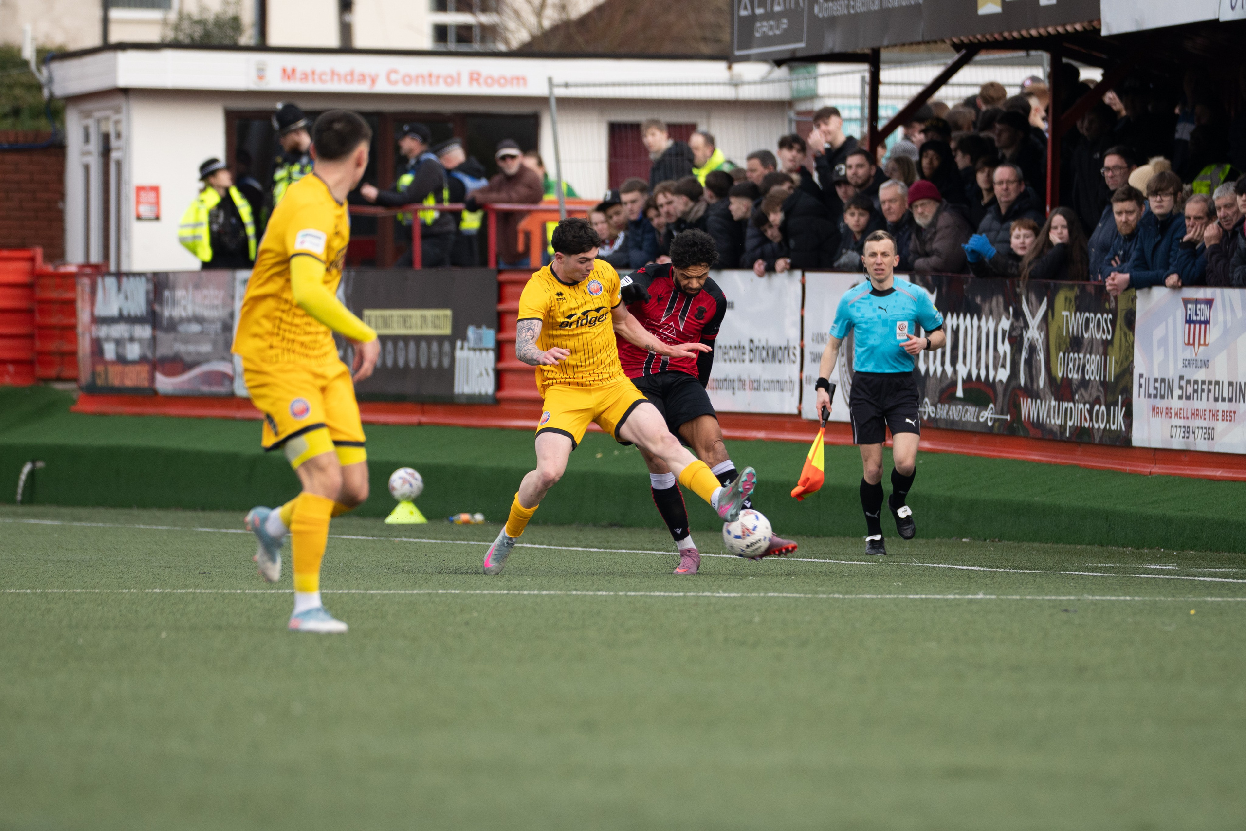 Tamworth, England — February 14, 2026: A Tamworth FC player challenges an Aldershot Town player for possession near the touchline as the assistant referee looks on during the Enterprise National League match at The Lamb Ground. Photo: Jay Soundo